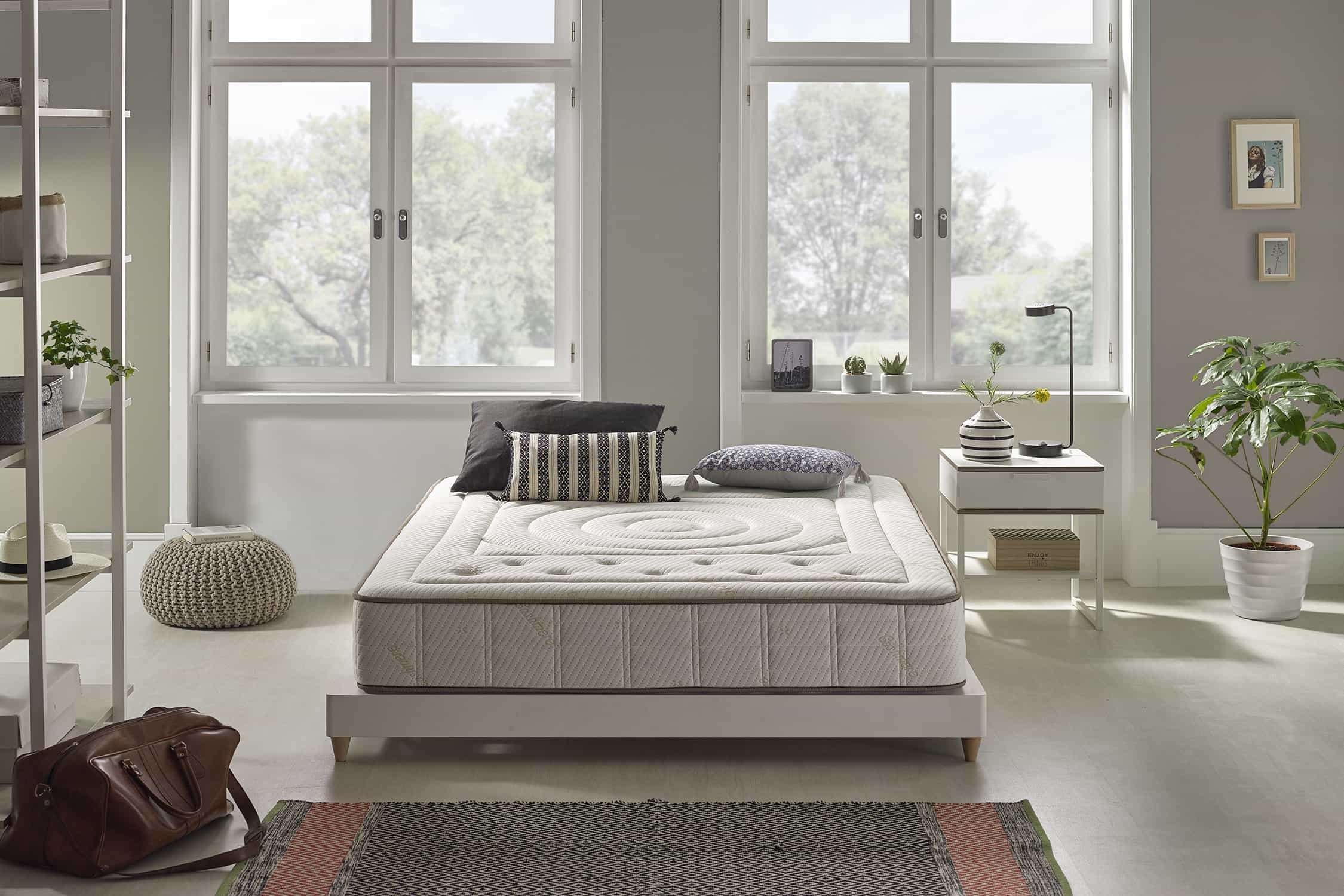 Escena de dormitorio con cama, ventanas y tonos neutros. Colchón en marco blanco.