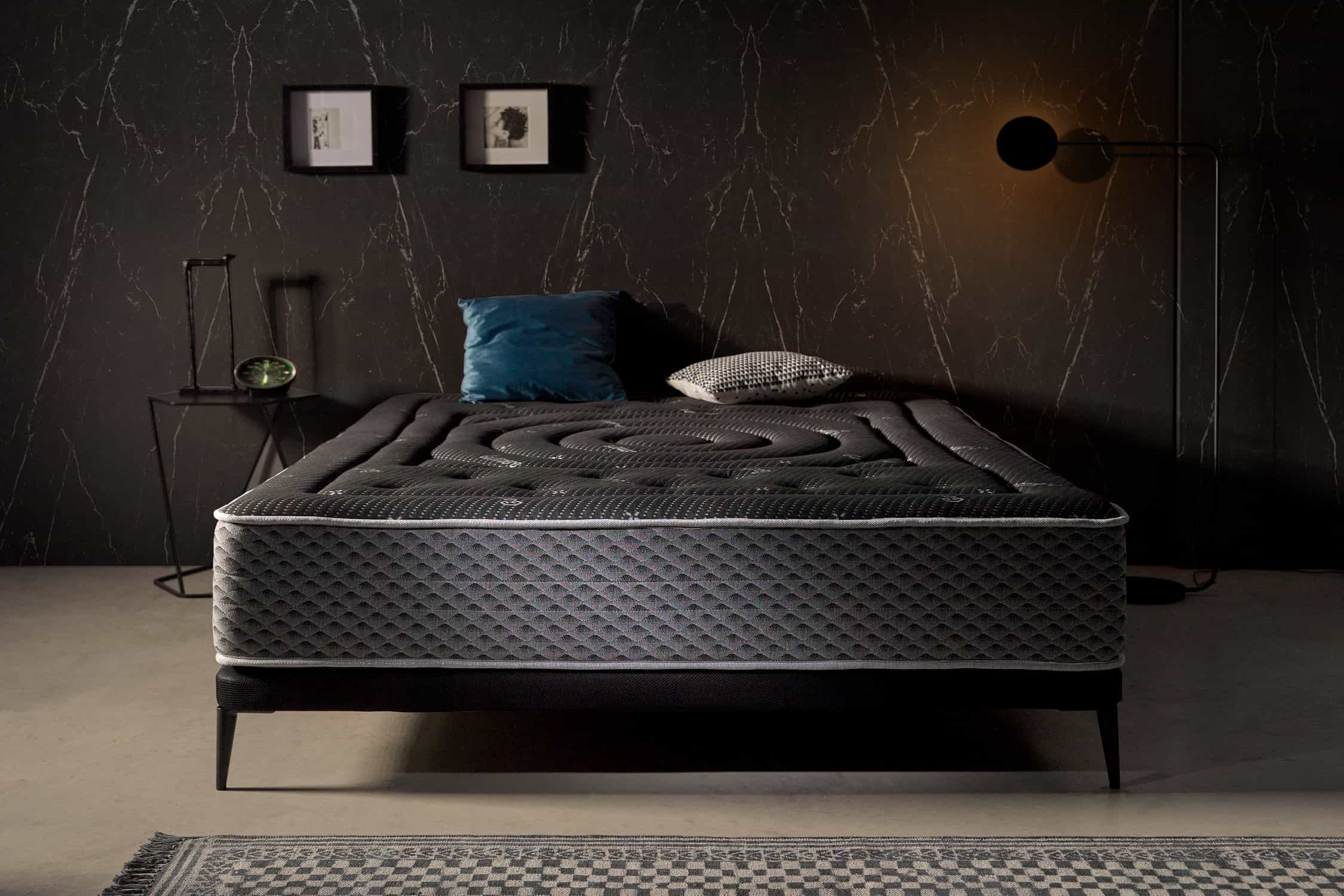 Escena de dormitorio con colchón gris oscuro, pared negra y decoración moderna.