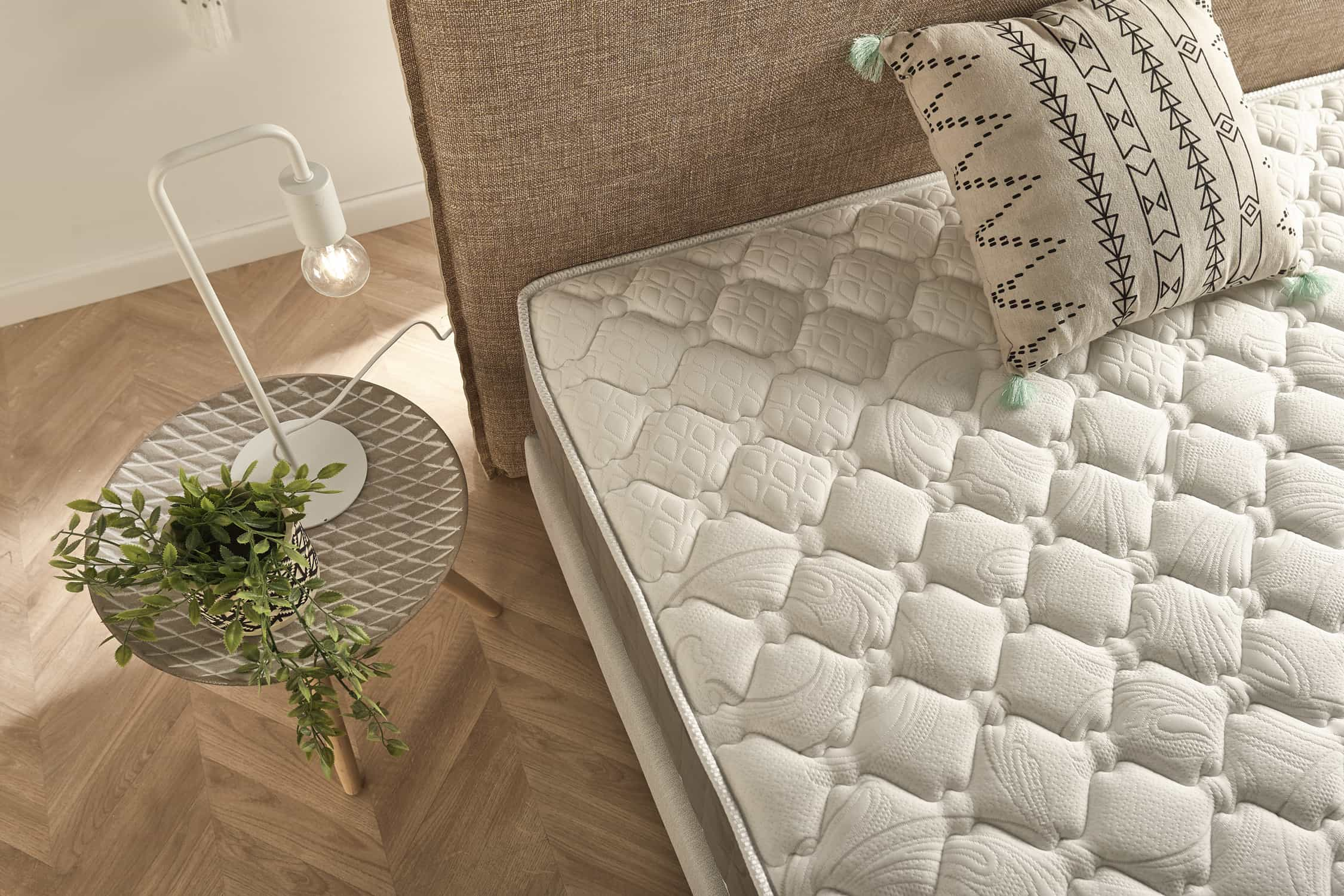Vista aérea de una cama con colchón blanco, mesita auxiliar con lámpara y suelo de madera.