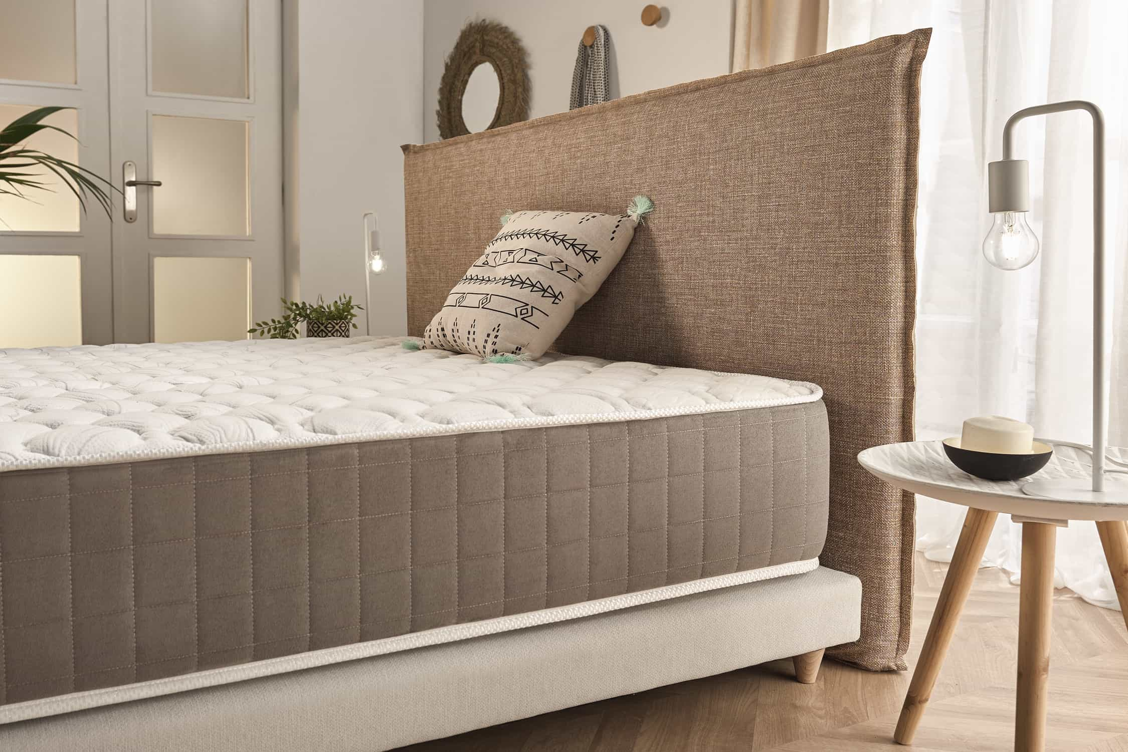 Una cama con cabecero beige, un colchón y una mesita auxiliar con una lámpara y un adorno.