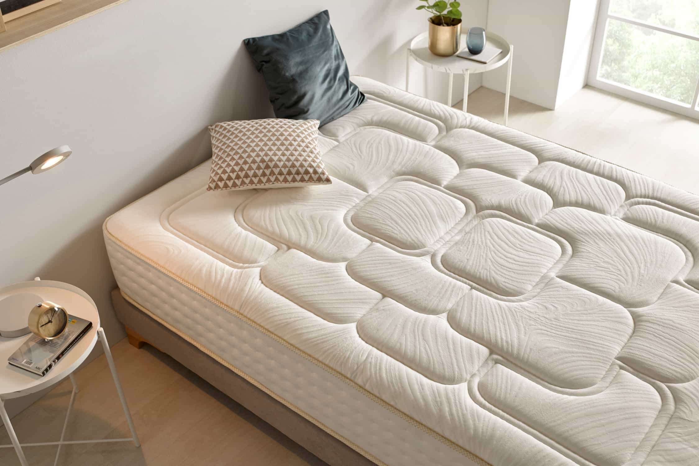 Una cama con colchón blanco, almohadas y una mesita auxiliar con una lámpara y un adorno.