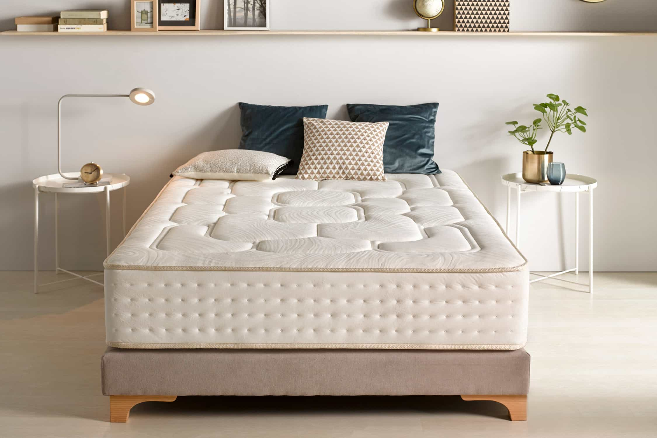 Una cama con colchón blanco, almohadas y una mesita auxiliar con una lámpara y una planta.