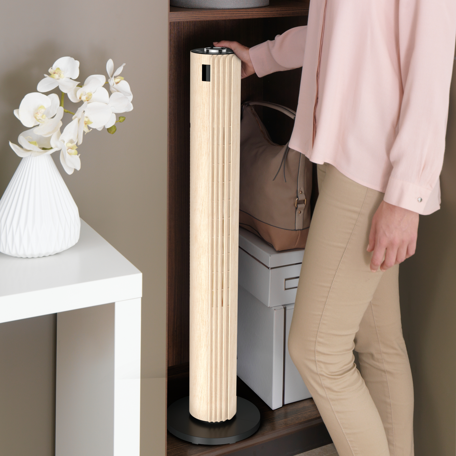 Kobieta dotyka beżowego wentylatora. Obok biały stół z kwiatami. W tle torba i pudełka.