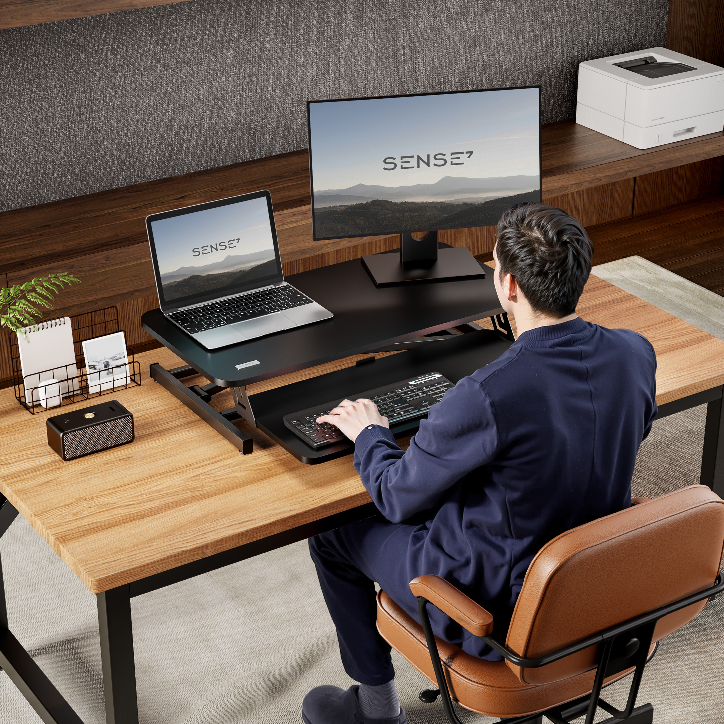 Mann, der an einem Schreibtisch mit Laptop und Monitor arbeitet. Der Schreibtisch ist braun mit schwarzen Metallbeinen.