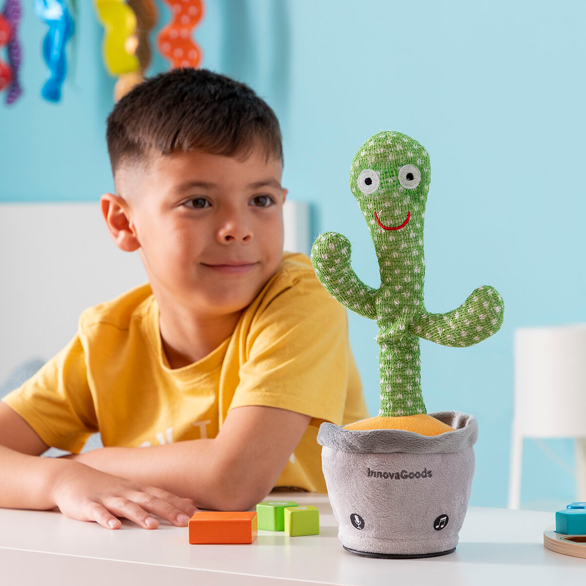 Un ragazzino accanto a un giocattolo a forma di cactus verde. Il giocattolo è in un vaso grigio.