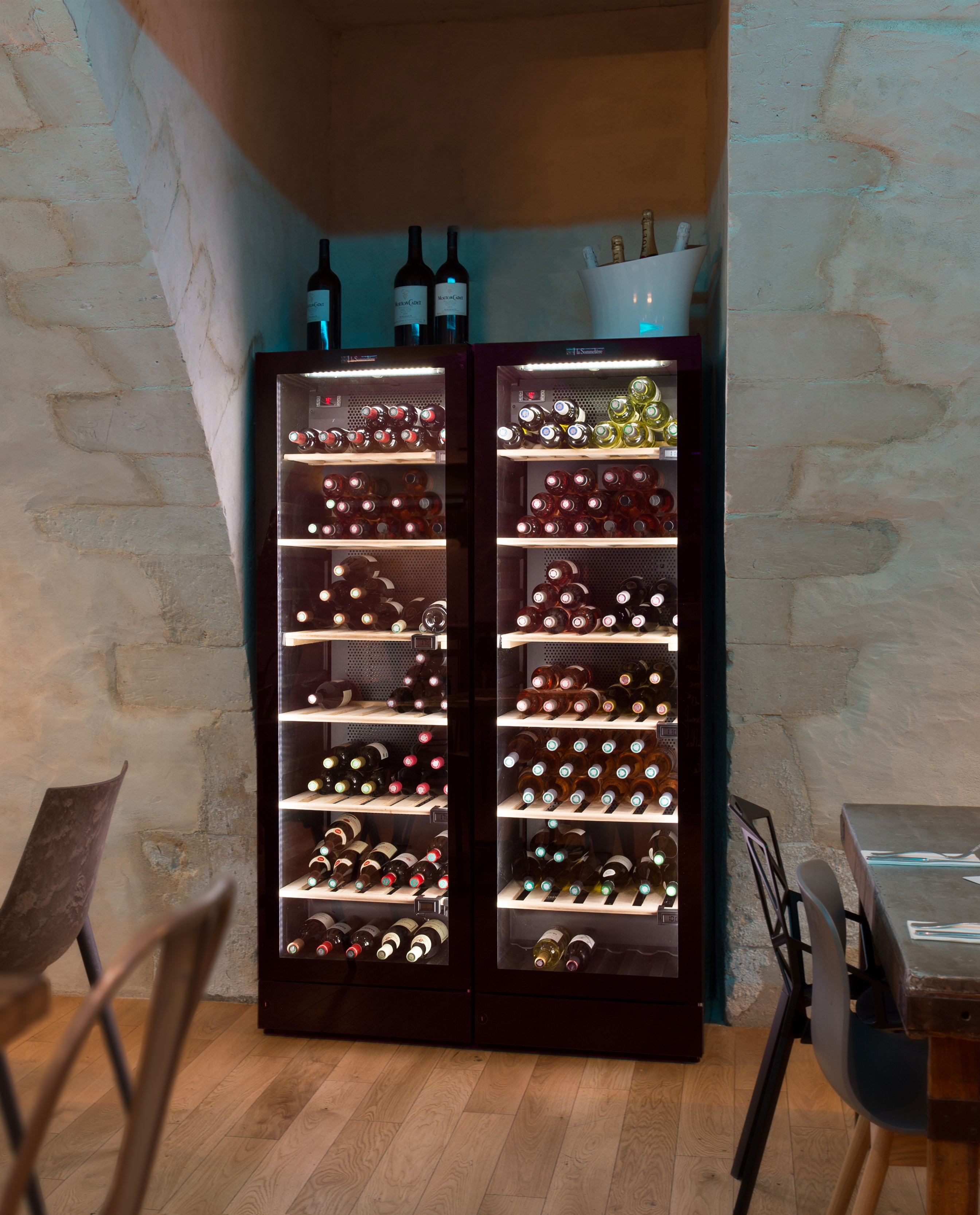 Dos enfriadores de vino con botellas en un restaurante. Detrás, pared de piedra y suelo de madera.