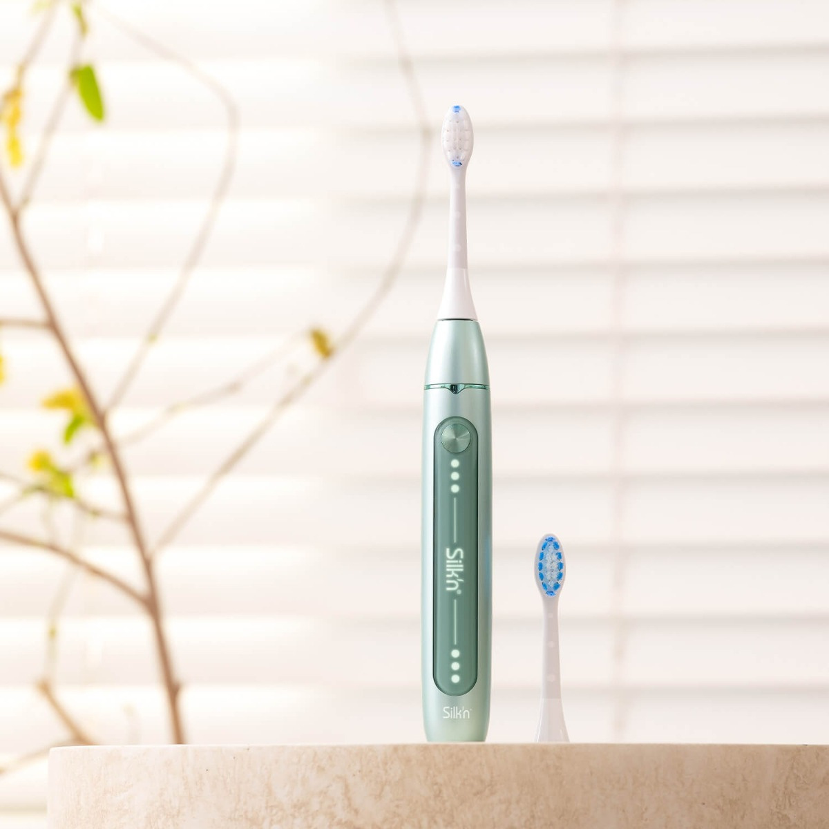 Zielona elektryczna szczoteczka do zębów i zapasowa główka na kamiennej powierzchni, z oknem w tle. Marka Silk'n.