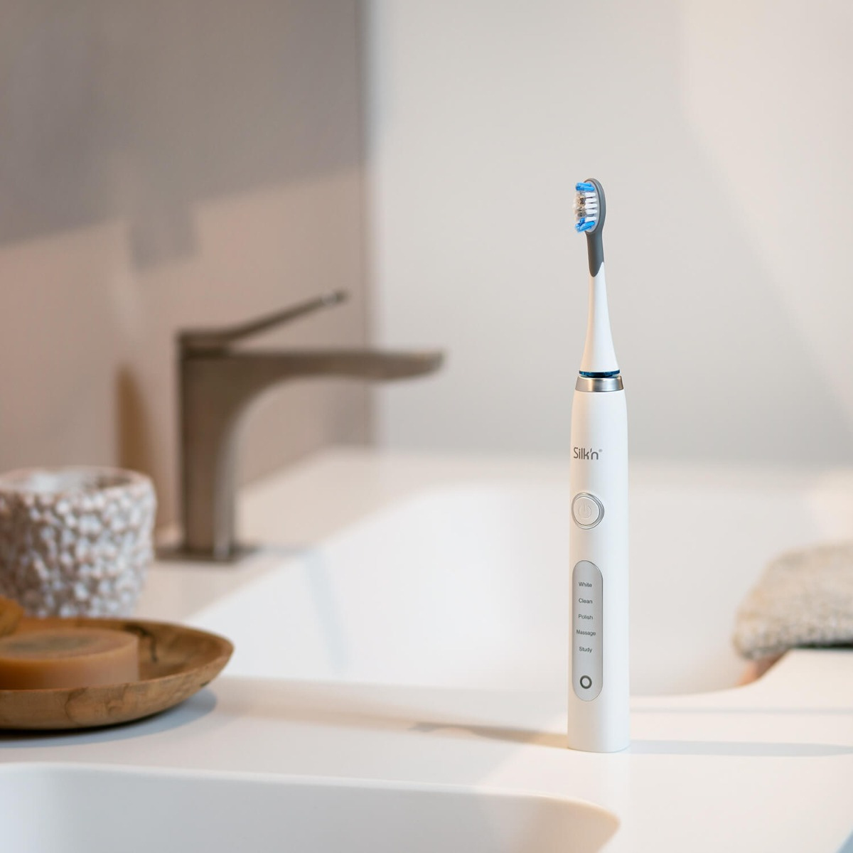 Biała szczoteczka do zębów Silk'n stojąca na białym zlewie, z kranem w tle.