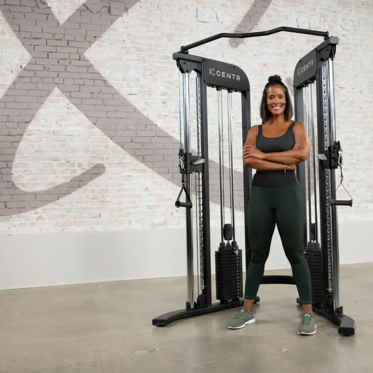 Femme debout dans une salle de sport, bras croisés, près d'une machine à câbles avec des poids ; fond de mur en briques.