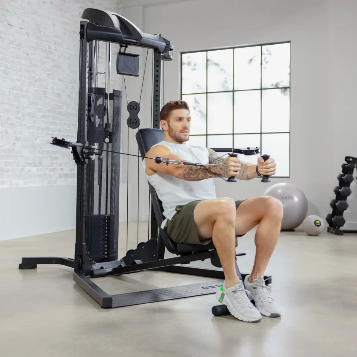 Mann benutzt eine Kabelmaschine, sitzend. Fitnessstudio, weiße Ziegelwand, Gewichte und ein großes Fenster.