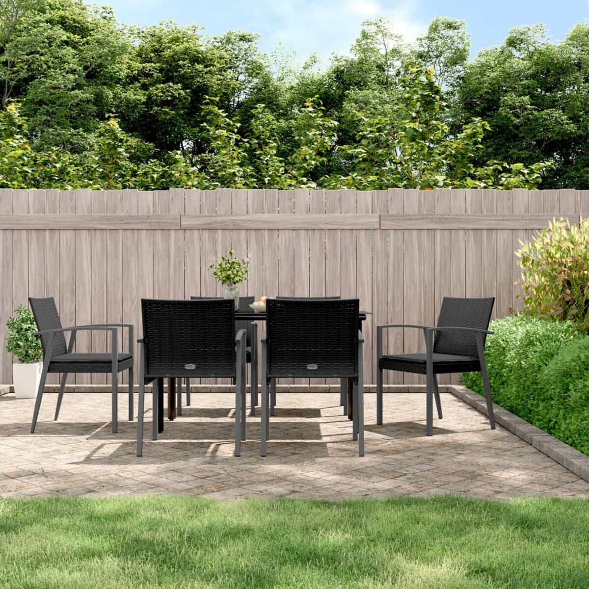 Szene auf einer Terrasse im Freien mit Tisch, Stühlen und Pflanzen. Ein Holzzaun und grüne Bäume im Hintergrund.