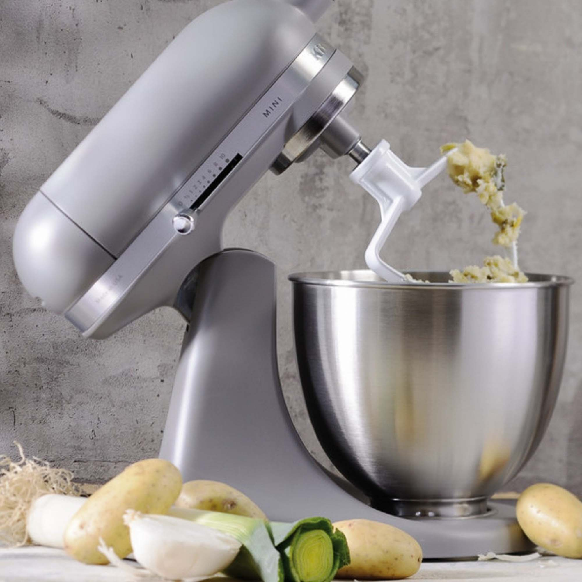 KitchenAid Mini Mixer. Grau, Rührschüssel mit Essen, Gemüse davor: Zwiebeln, Kartoffeln, Lauch.