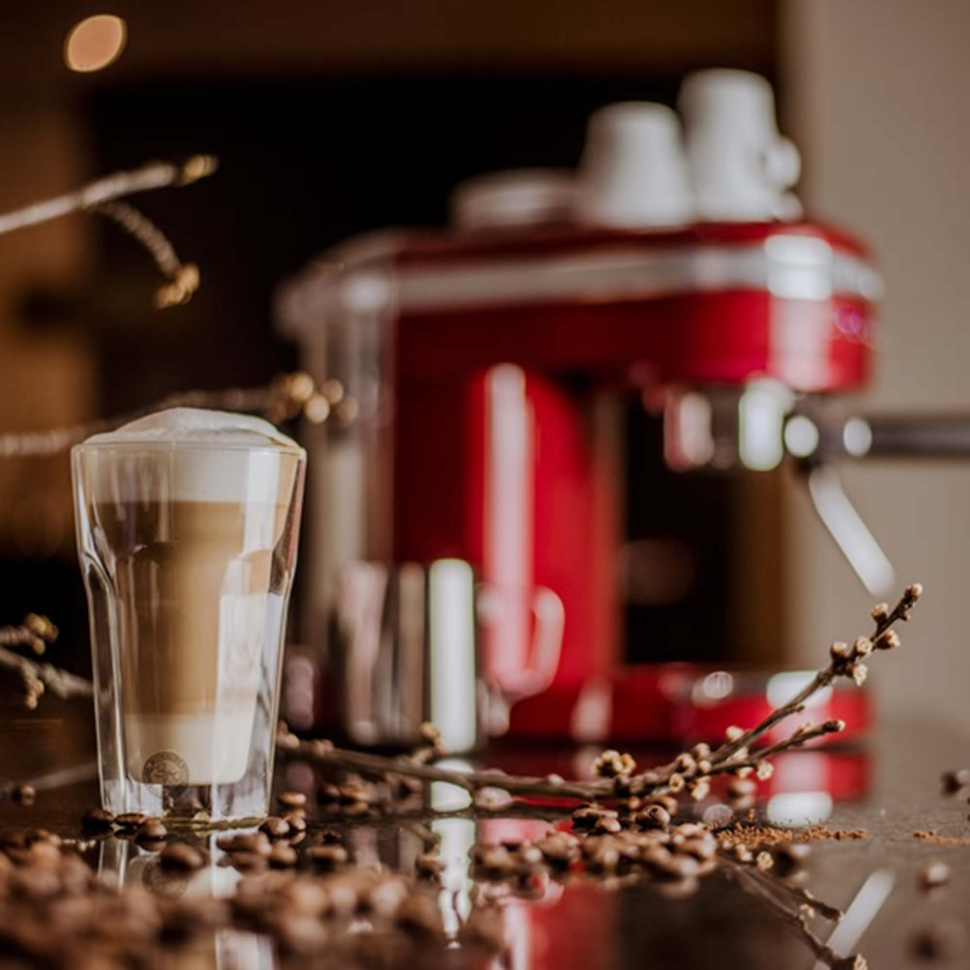 Latte in einem Glas mit Kaffeebohnen und Zweigen vor einer roten Espressomaschine.