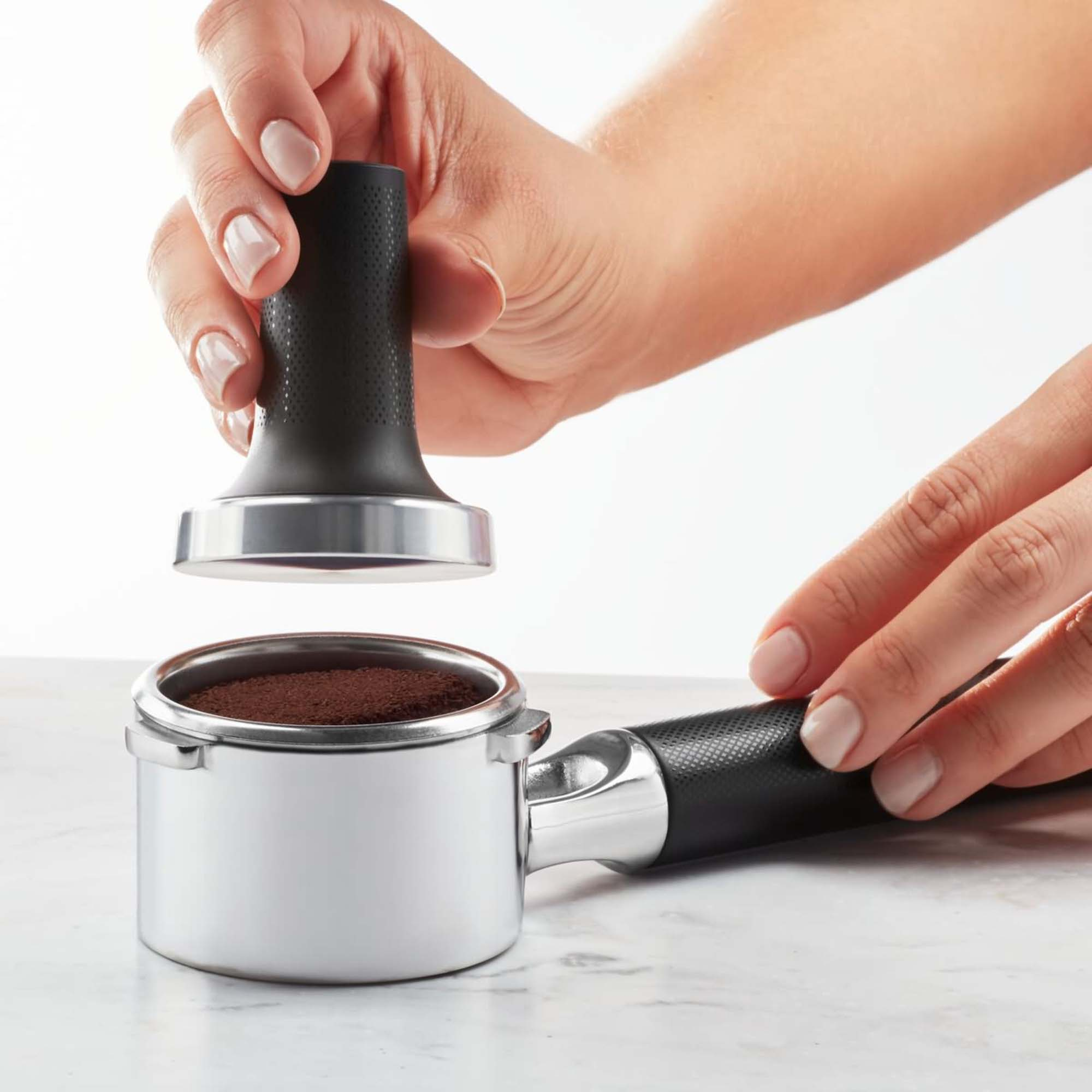 Hände, die Kaffeepulver mit einem Tamper in einen Siebträger pressen, vor weißem Hintergrund.