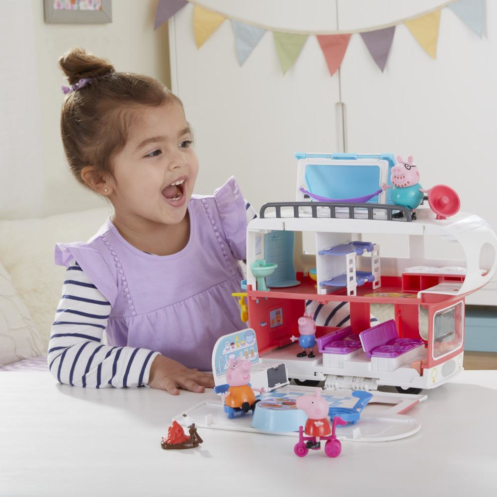 Ein junges Mädchen lächelt und spielt mit einem Peppa Wutz Wohnmobil-Spielset auf einem Tisch.