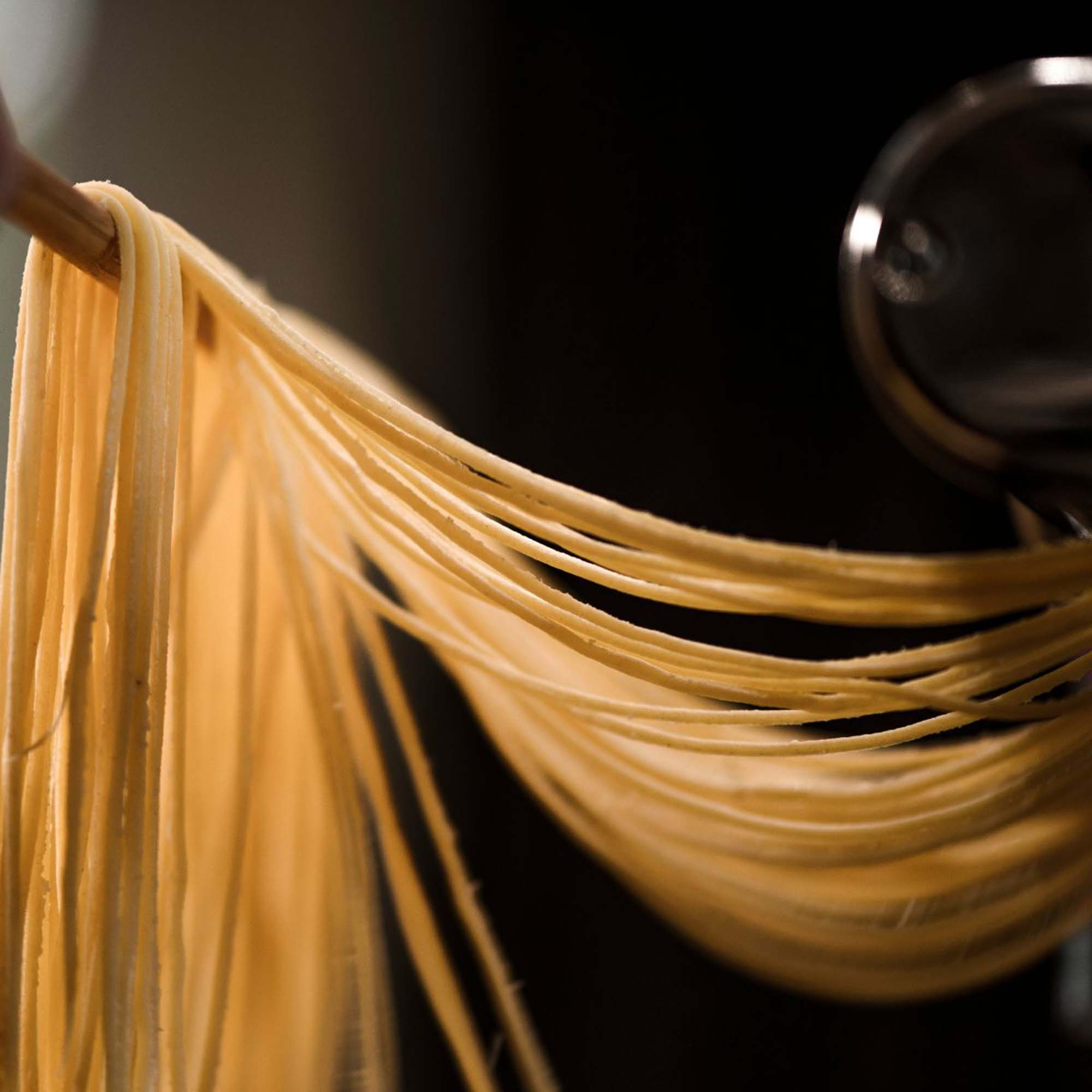 Hängende Spaghetti-Nudeln, gehalten von einem Holzwerkzeug, dunkler Hintergrund.