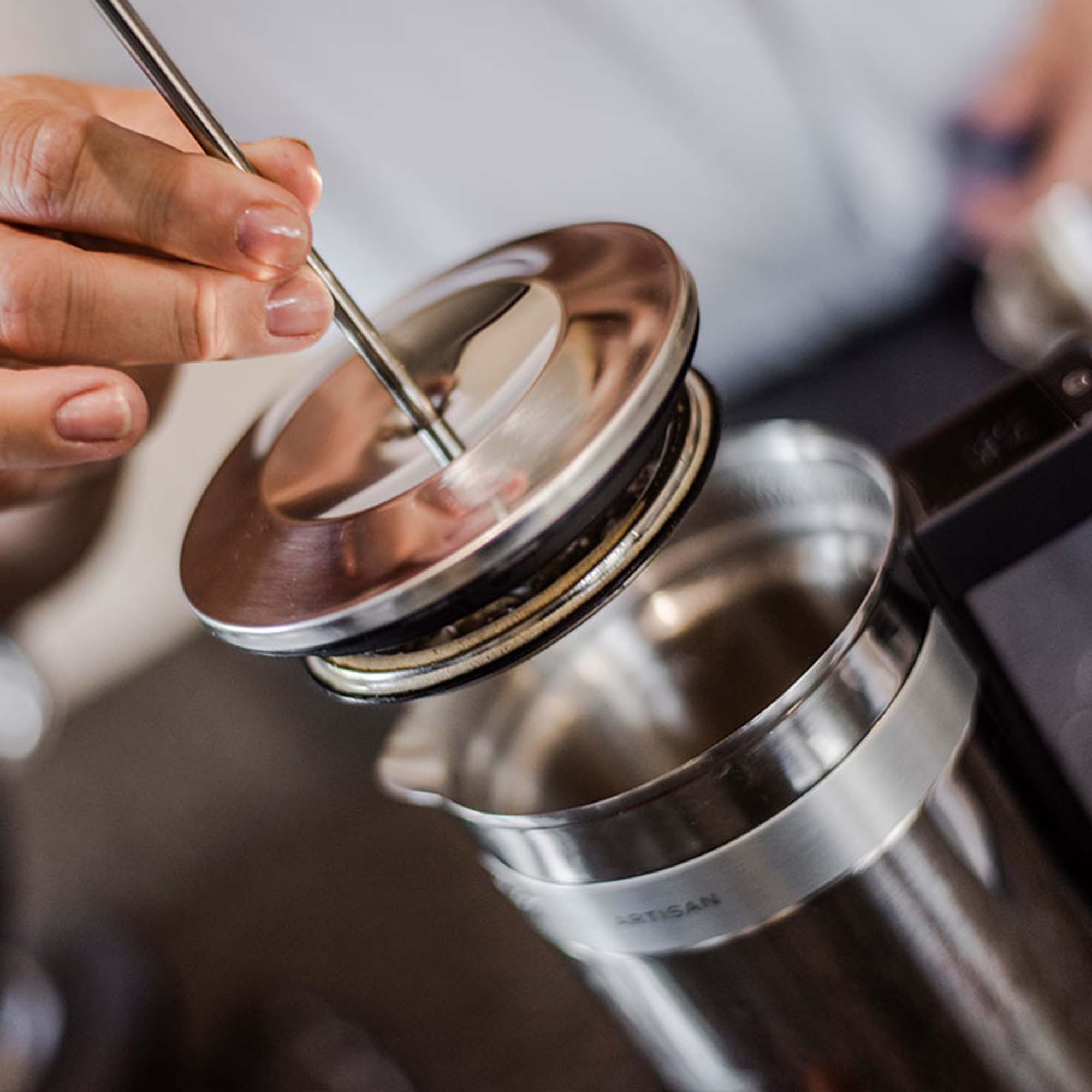 Hand öffnet den Deckel einer Kaffee-Kanne aus Edelstahl, mit Artisan-Etikett.