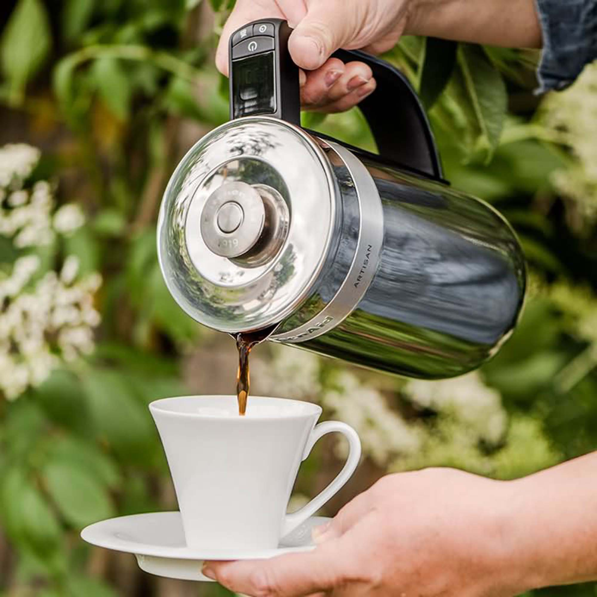Kaffee gießt aus einer silbernen Kaffeemaschine in eine weiße Tasse.
