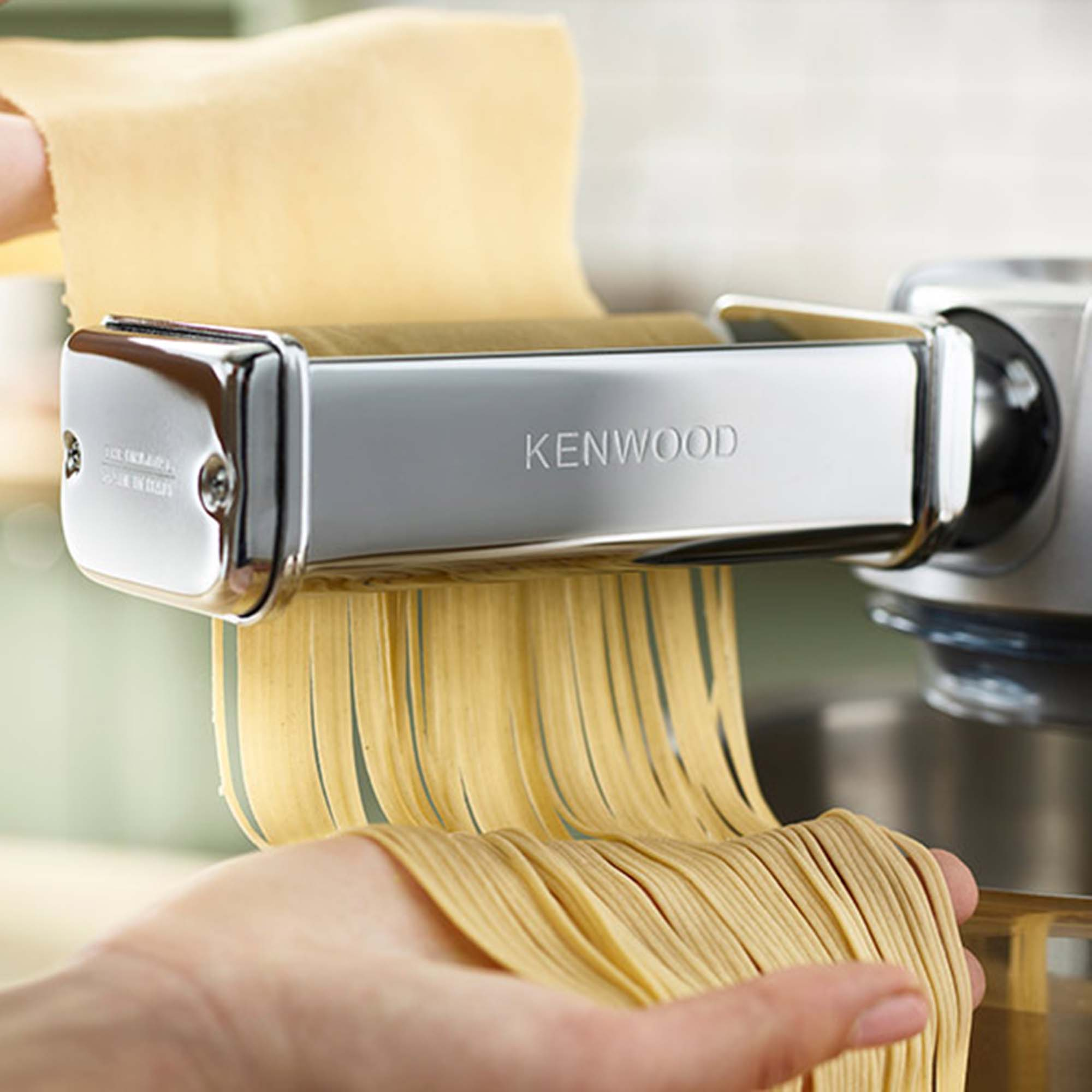 Maszyna do makaronu rozwałkowująca makaron. Makaron spada na dłoń. Kenwood to nazwa marki.