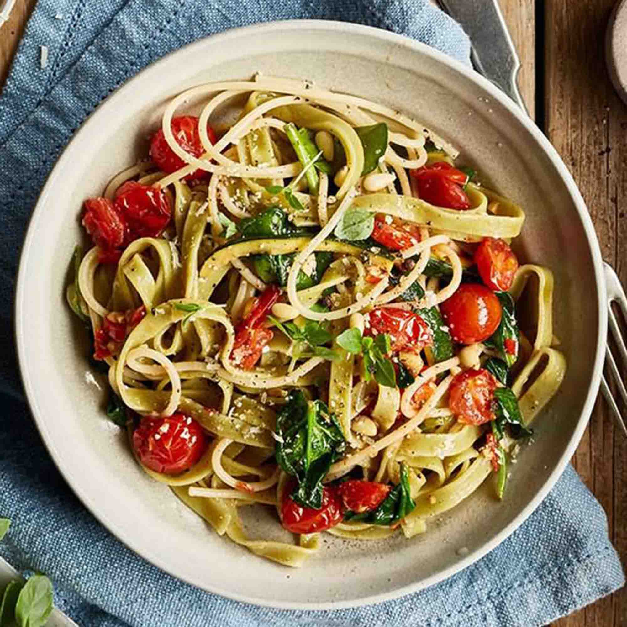 Danie z makaronem z pomidorami, szpinakiem, orzeszkami piniowymi i bazylią. Drewniany stół i niebieska serweta w tle.