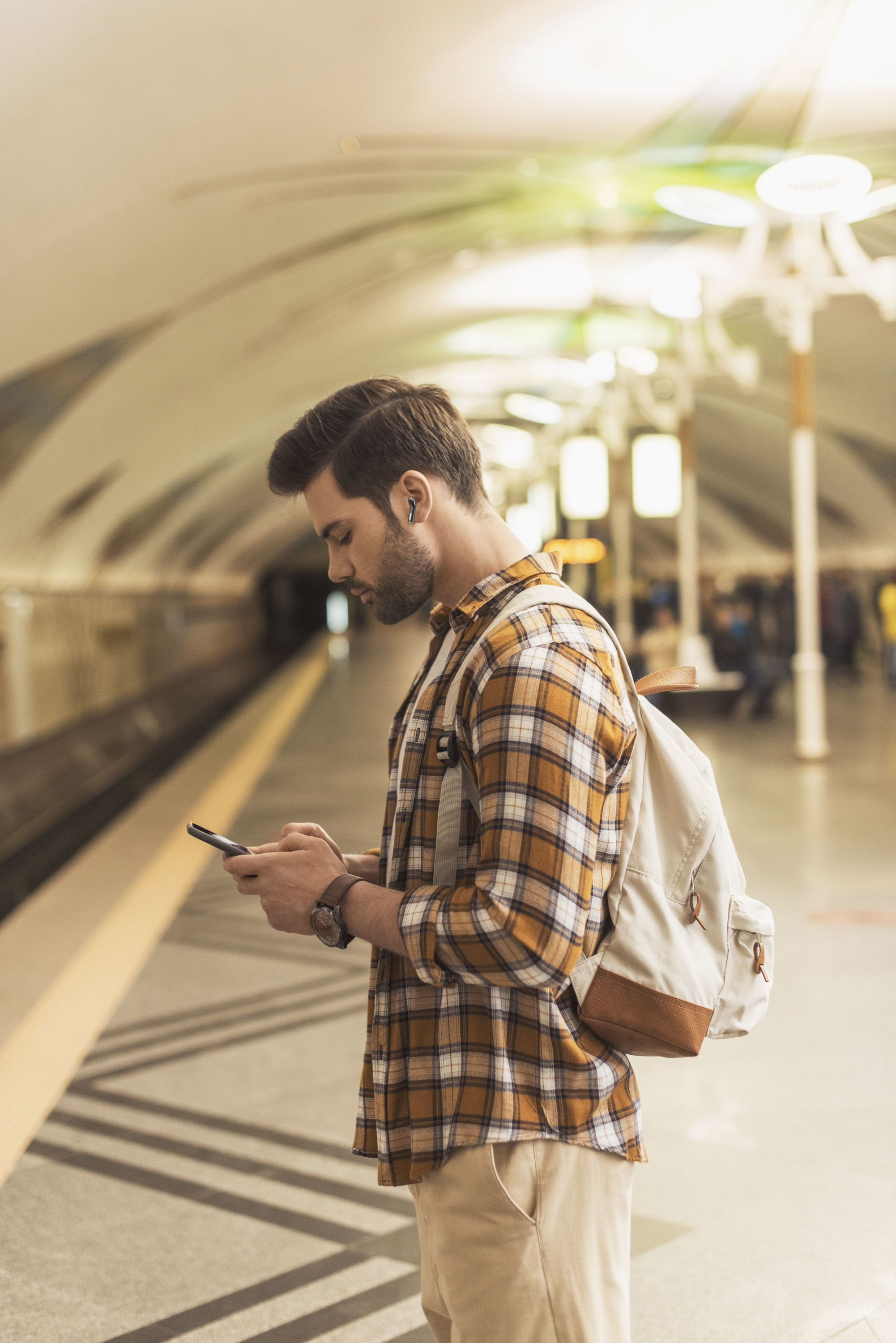Mann in einer U-Bahn-Station, der auf sein Handy schaut, mit Ohrhörern und Rucksack.