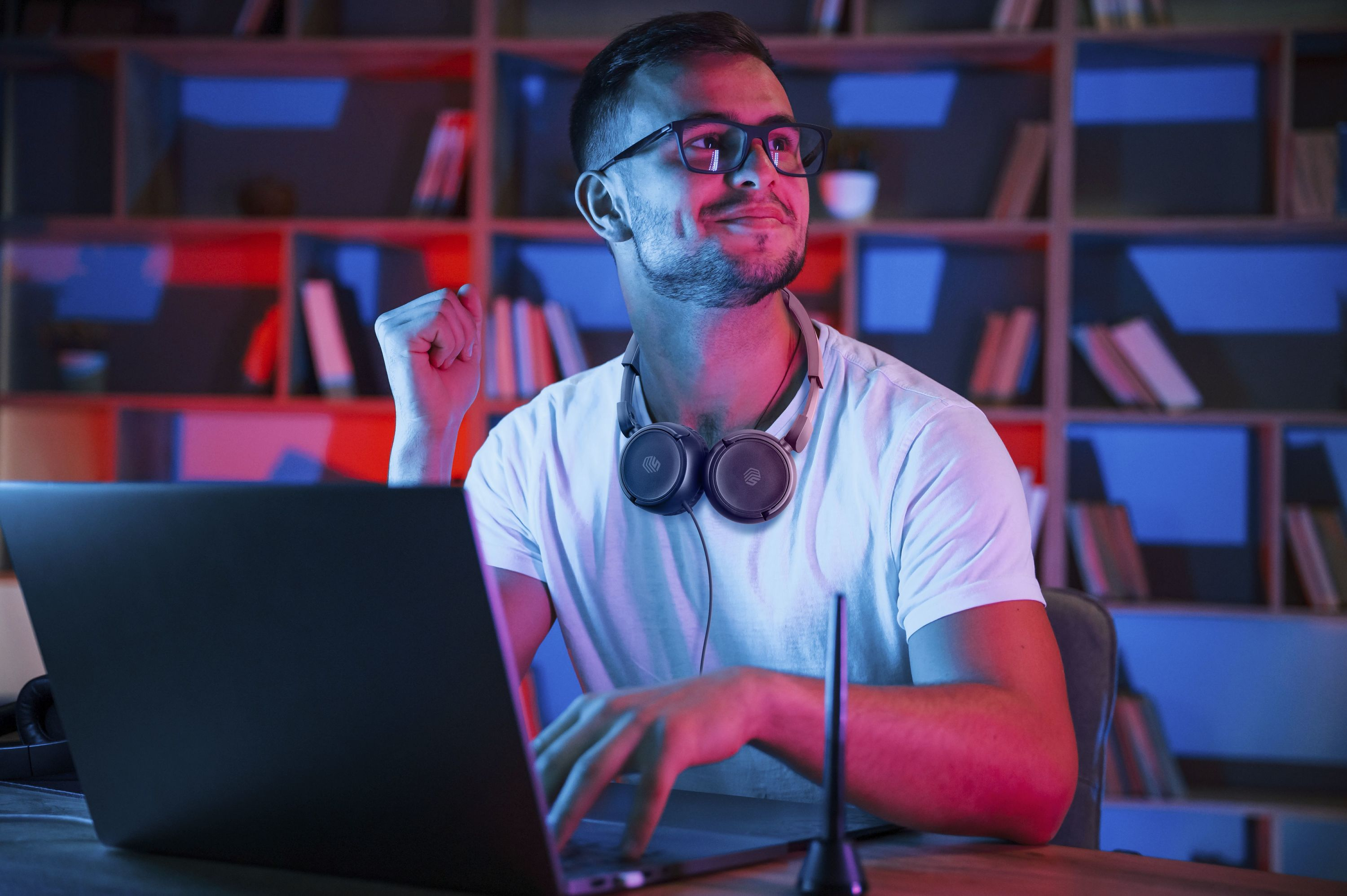 Uomo con gli occhiali che usa un laptop; cuffie al collo, che guarda in alto. Libreria sullo sfondo.