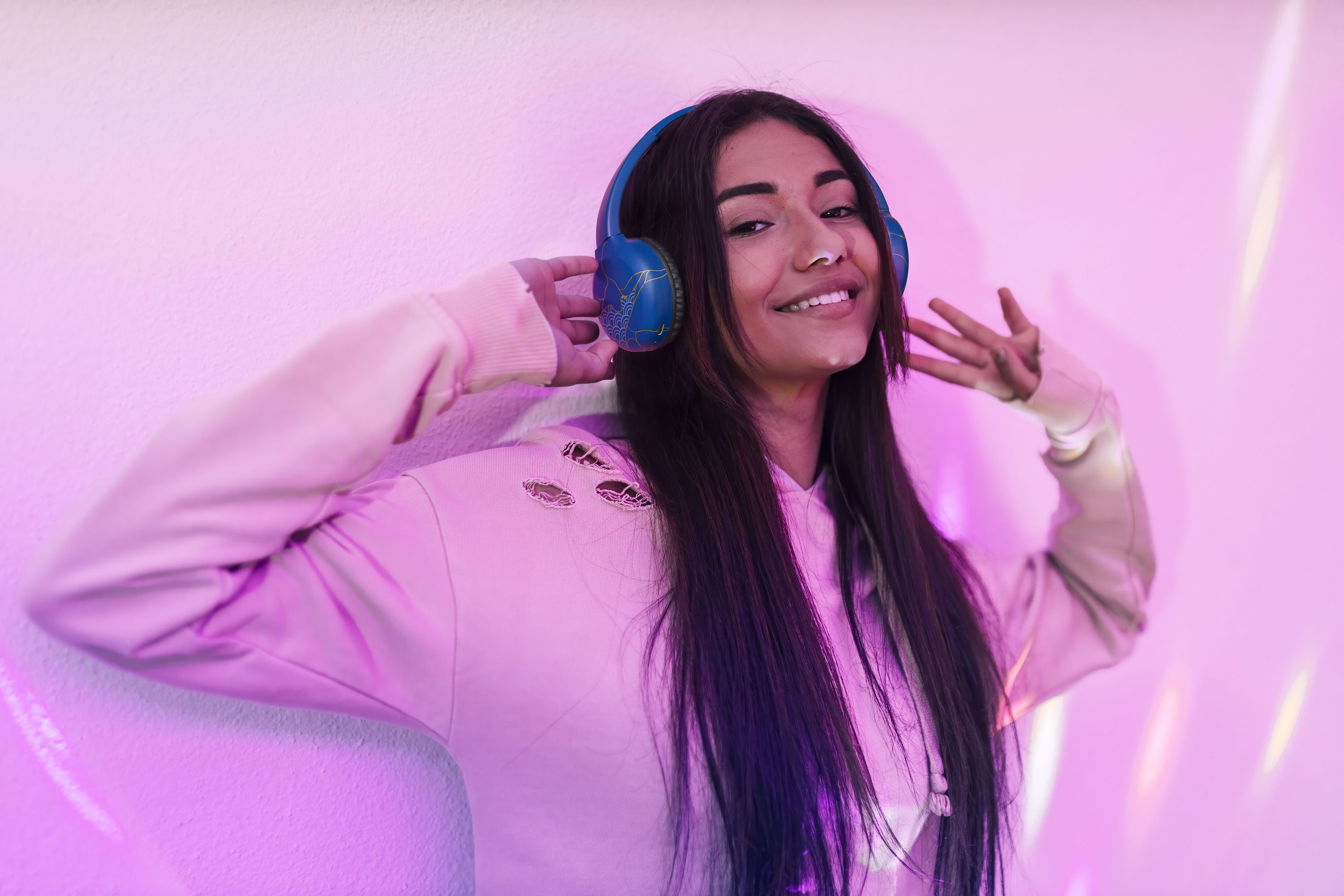 Donna con cuffie blu, sorridente. Parete rosa con effetti di luce al neon rosa e viola.