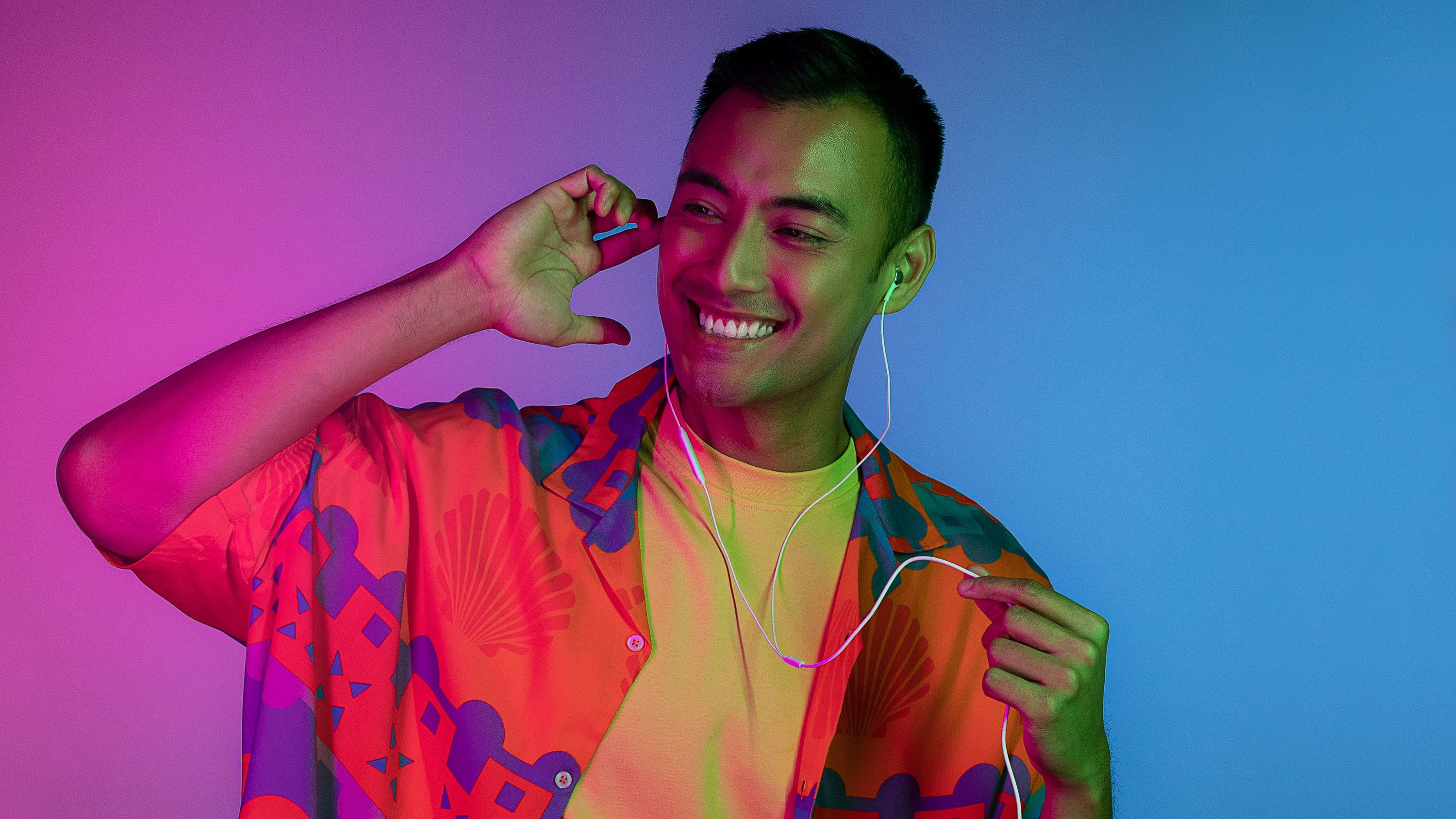 Mann mit orangefarbenem Hemd und Kopfhörern. Lächelt, hört Musik. Rosa-blauer Hintergrund.