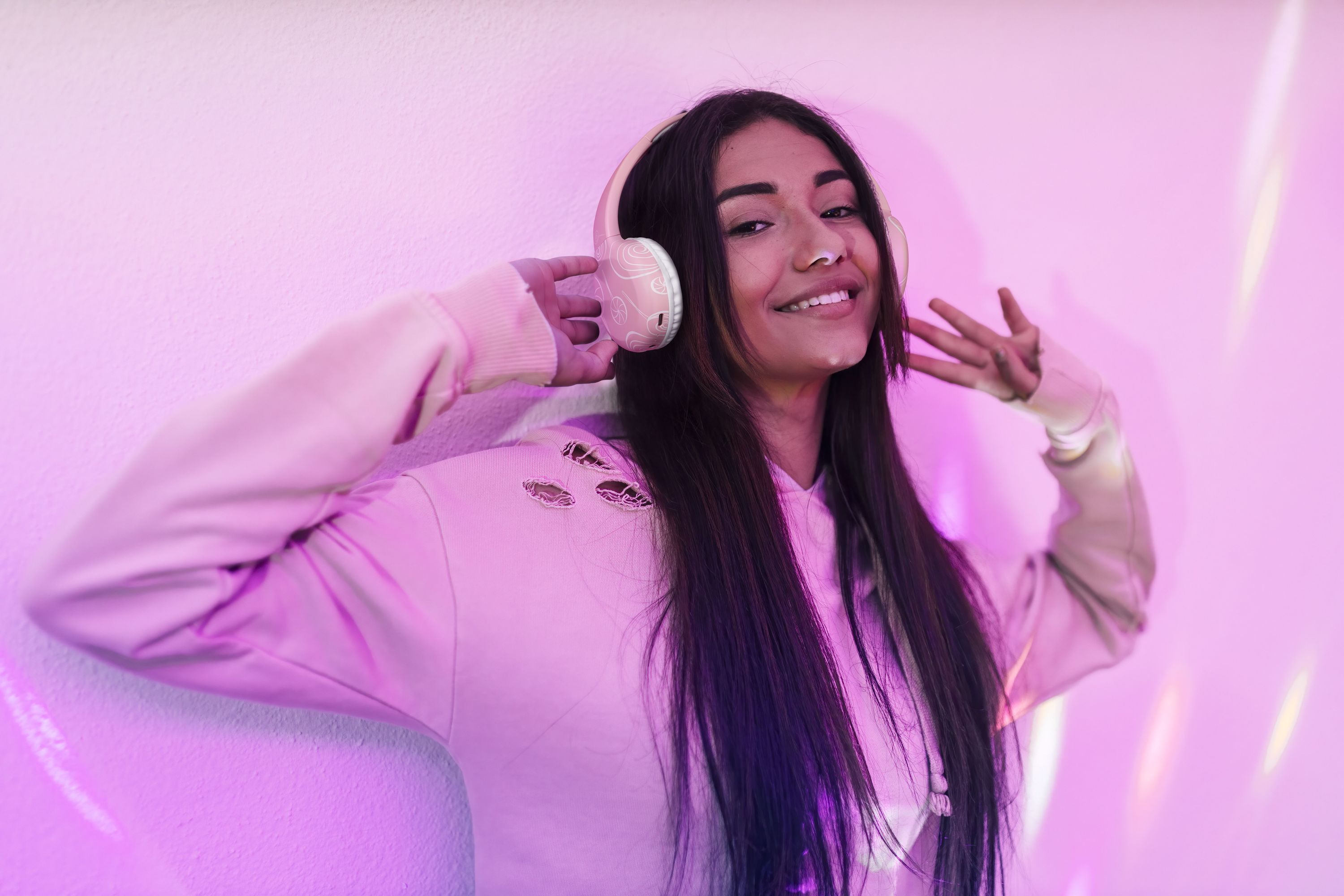 Młoda kobieta uśmiechnięta, w słuchawkach, oparta o różową ścianę z różowymi i fioletowymi światłami.