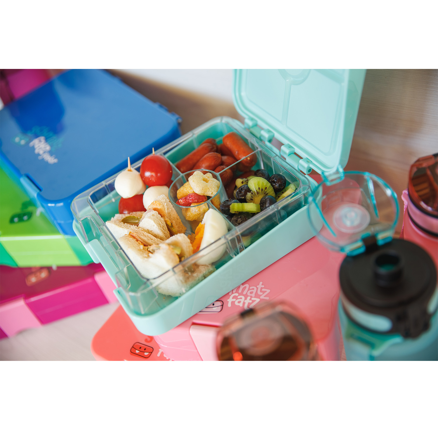 Eine Lunchbox mit Fächern, gefüllt mit Sandwich-Dreiecken, Obst und Snacks. Sie steht auf einer rosa Oberfläche.