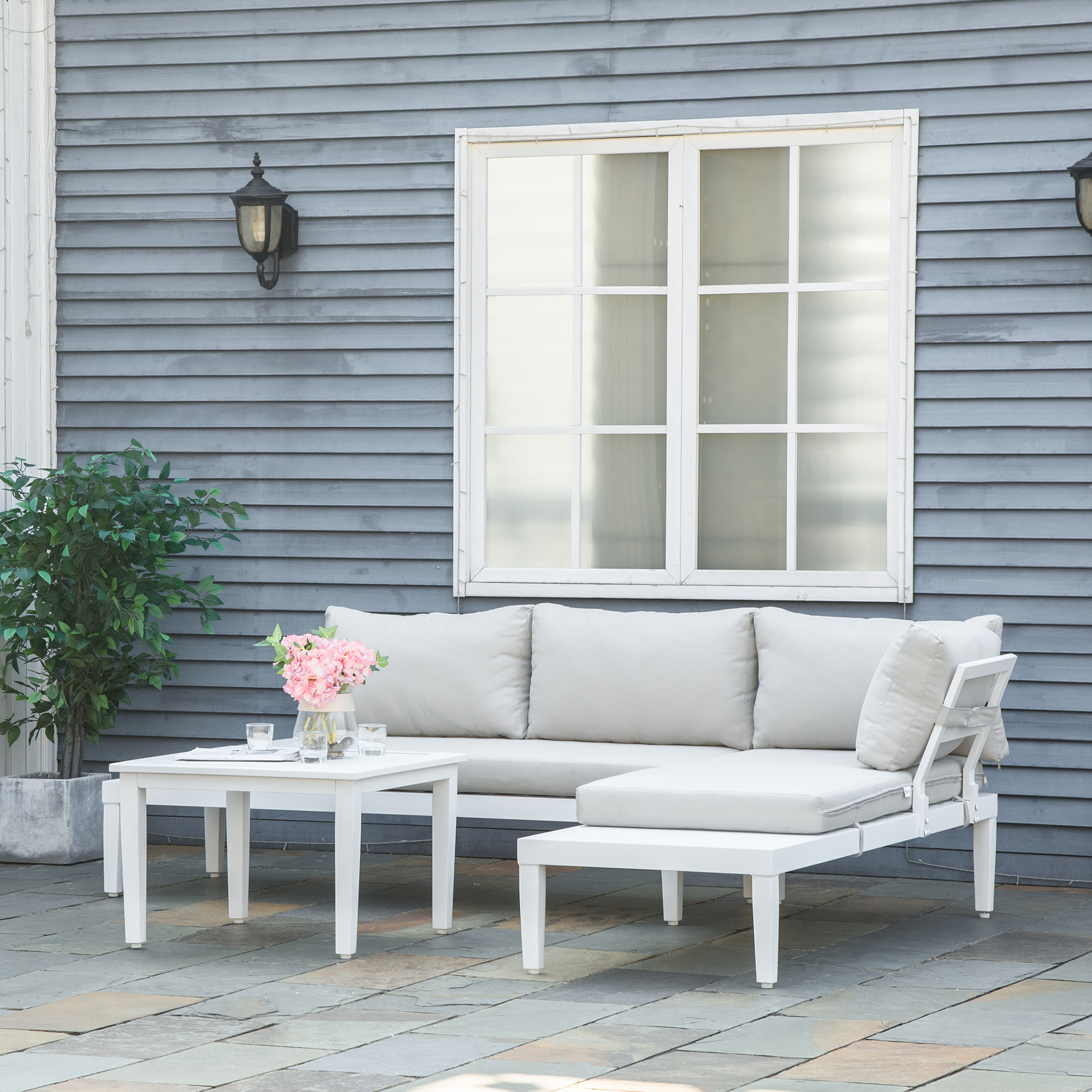 Ein weißes Outdoor-Ecksofa mit Kissen und einem kleinen Tisch mit rosa Blumen und Gläsern.