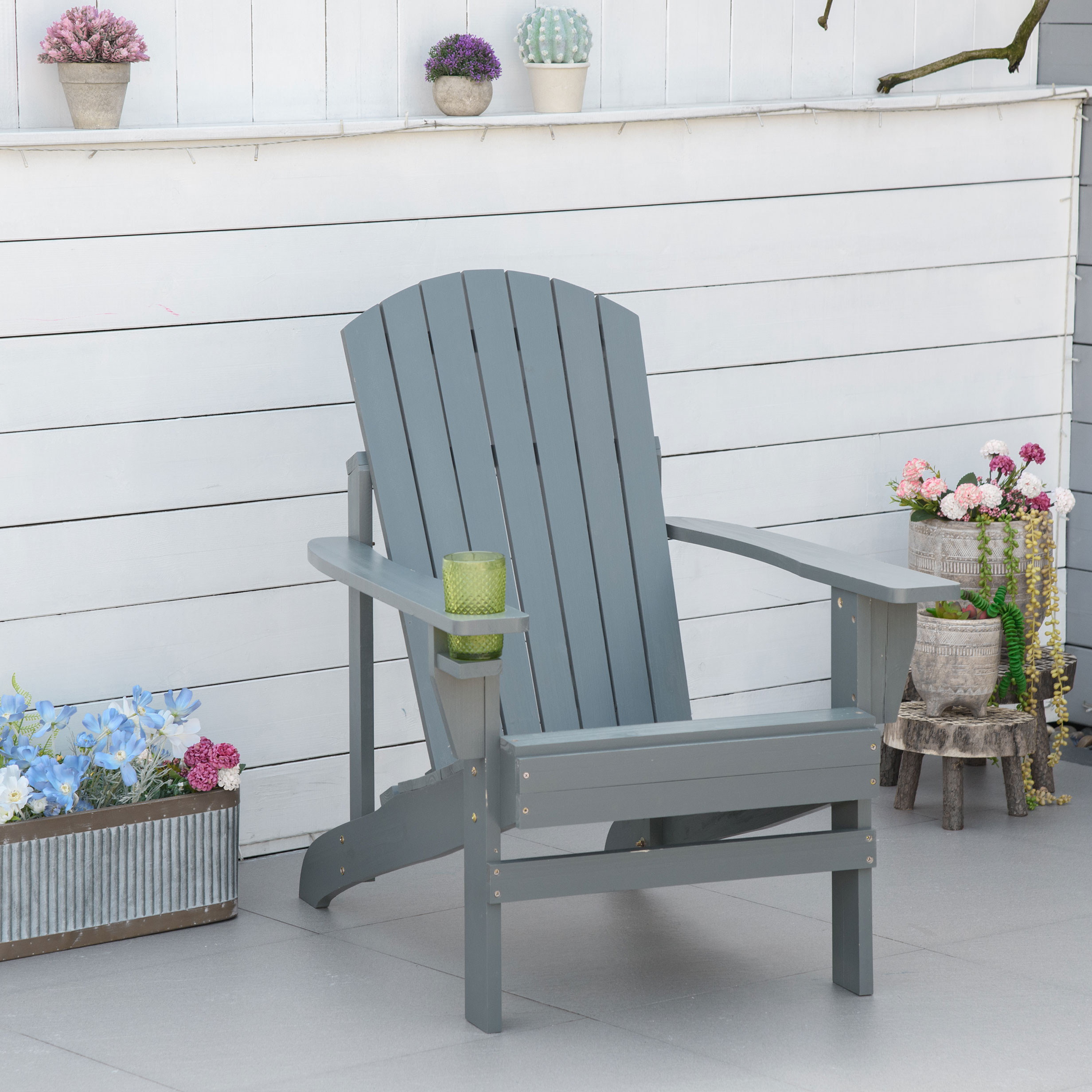 Grauer Adirondack-Stuhl aus Holz mit einem Glas in der Armlehne. Blumen und Pflanzen sind in der Nähe.