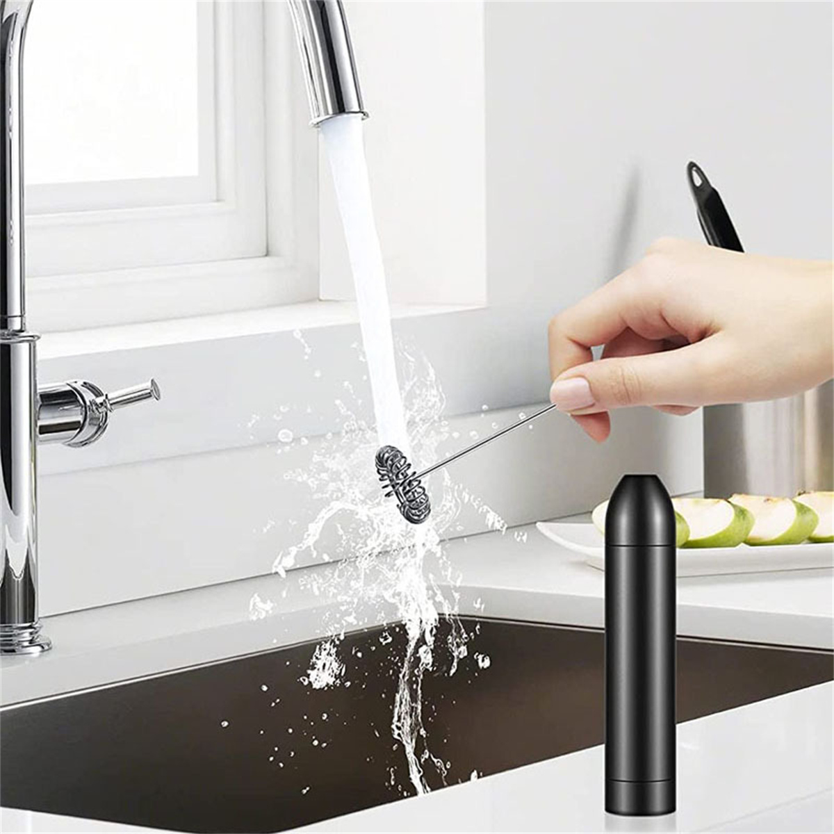 Eine Hand schäumt Milch mit einem Stab in einem Waschbecken, mit spritzendem Wasser. Äpfel sind auf der Theke.