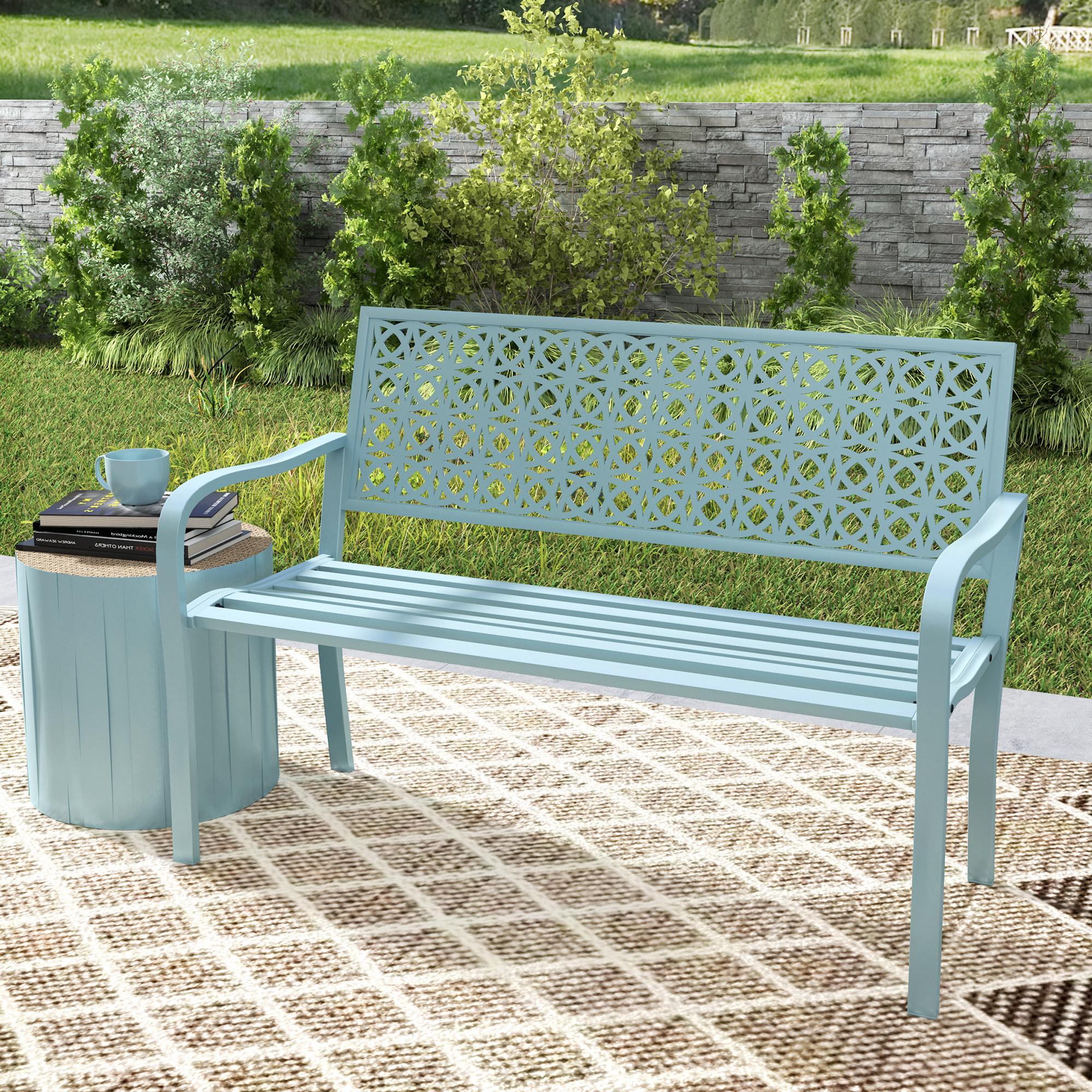 Eine hellblaue Metallbank und ein kleiner Tisch auf einem gemusterten Outdoor-Teppich, mit Grün im Hintergrund.