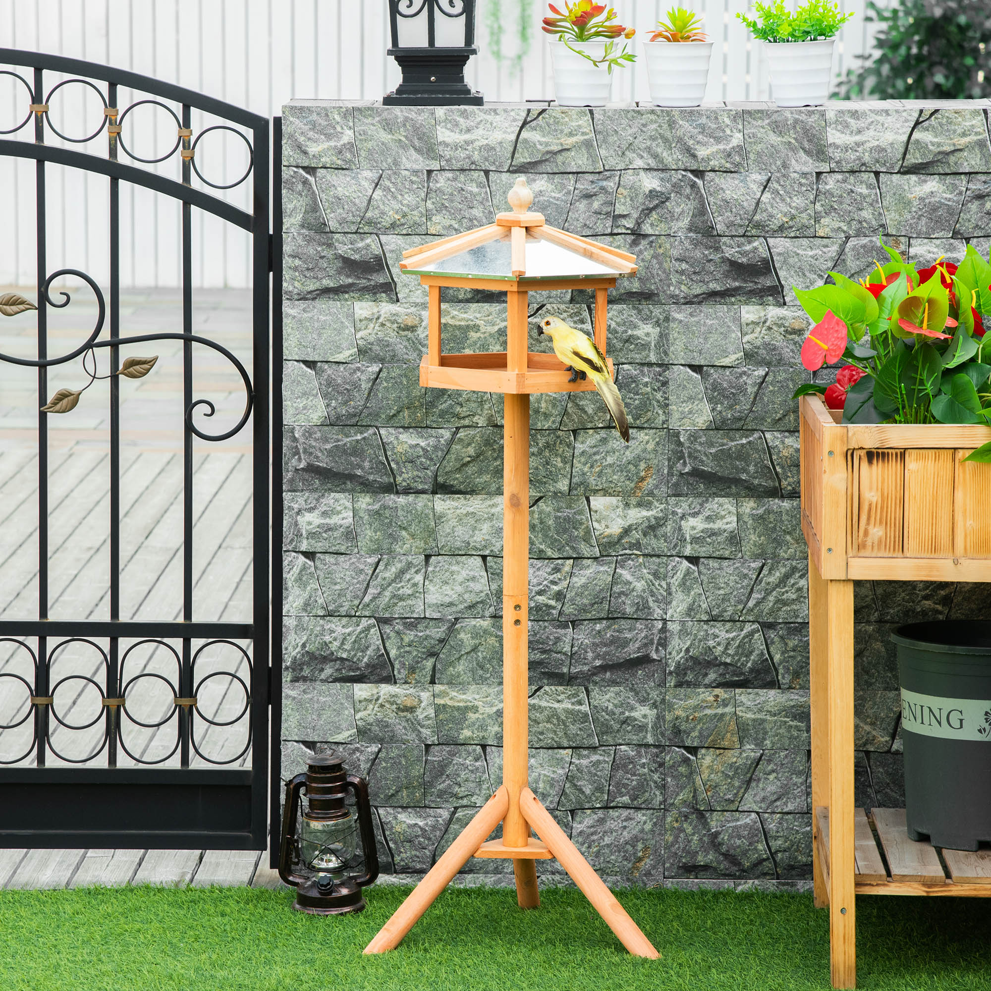 Vogelfutterautomat mit gelbem Vogel. Im Garten mit Steinmauer und schwarzem Tor.