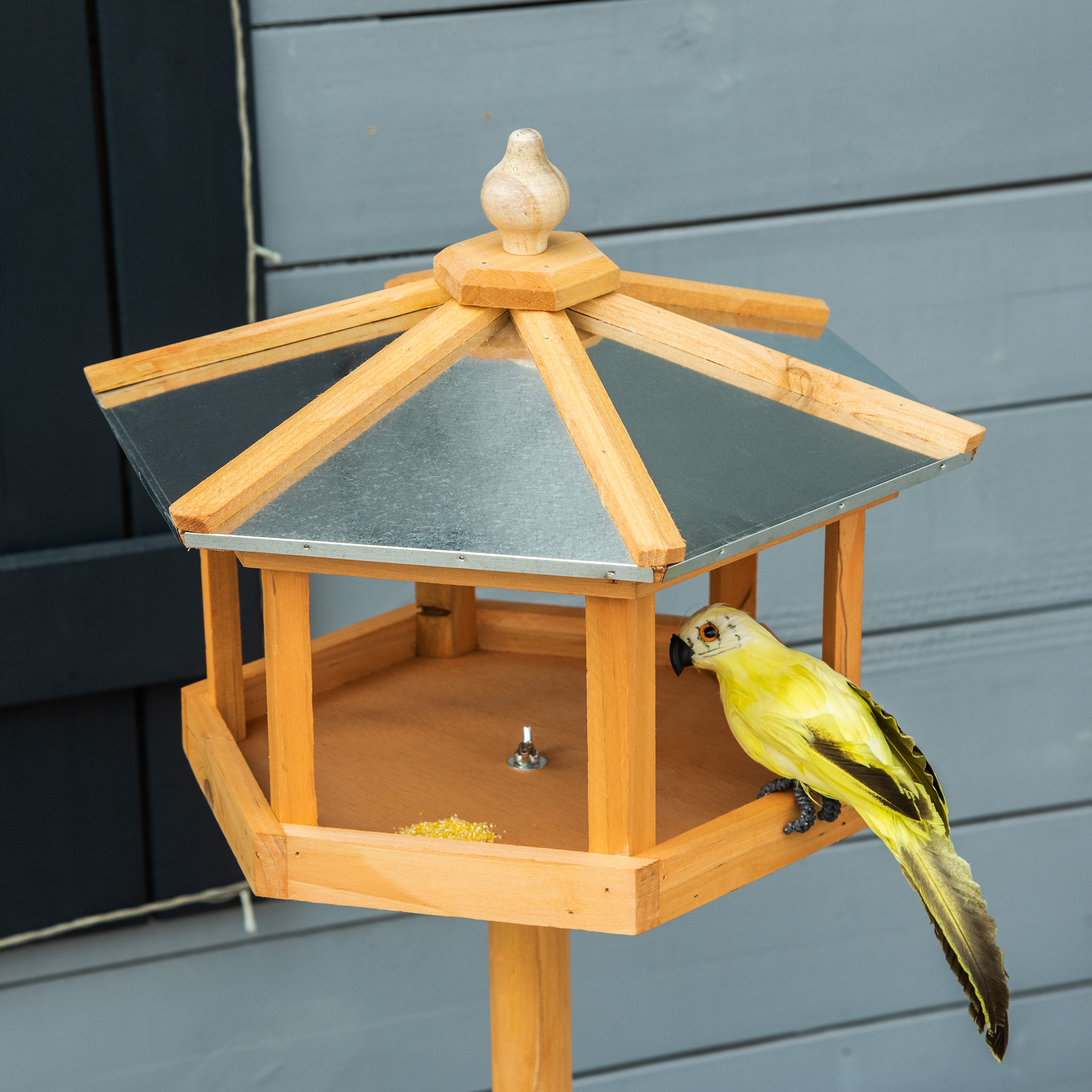 Vogelfutterautomat aus Holz mit gelbem Vogel. Nahaufnahme mit Details. Grauer Hintergrund.