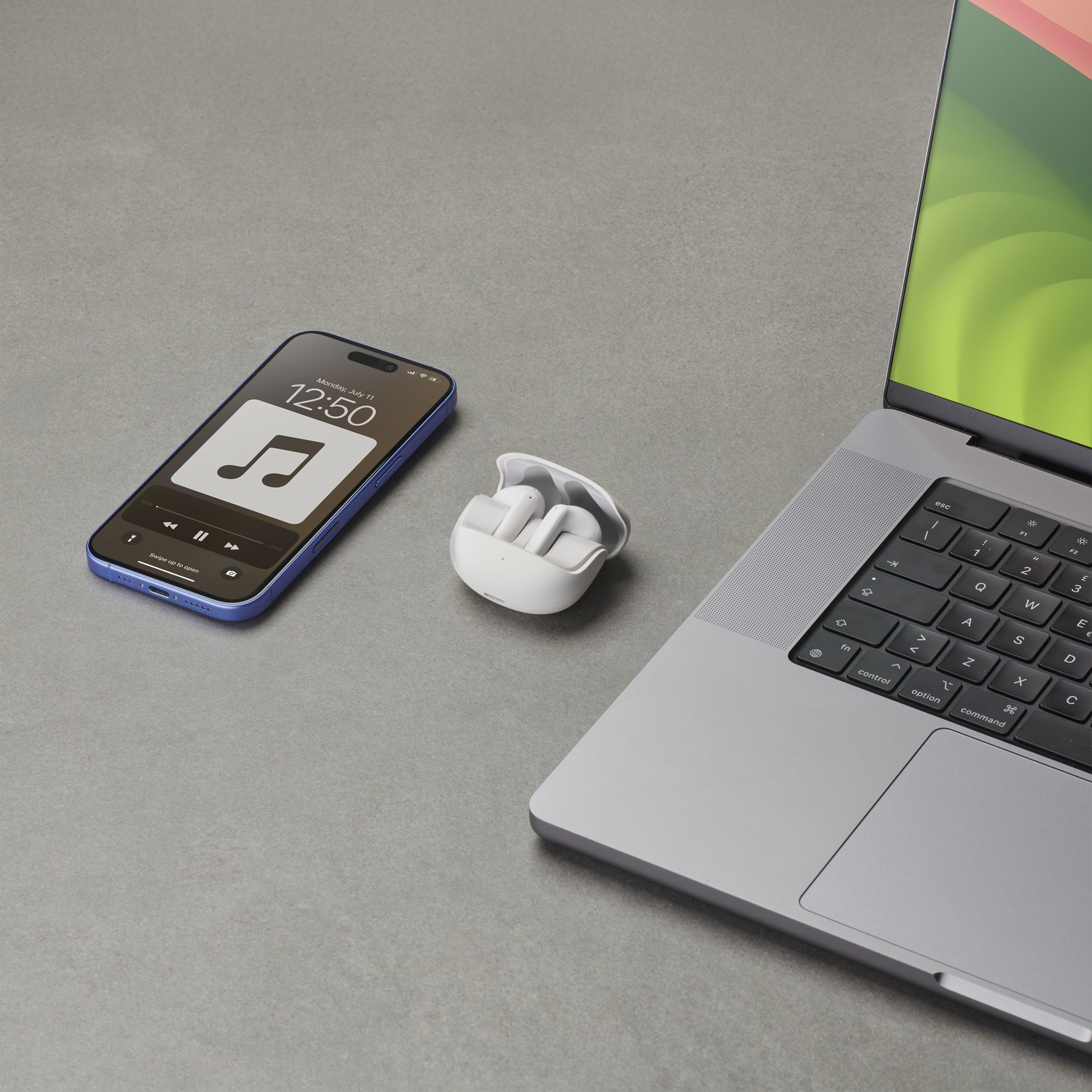 Auriculares blancos, estuche, un teléfono y un portátil sobre una superficie gris. El teléfono muestra una app de música.