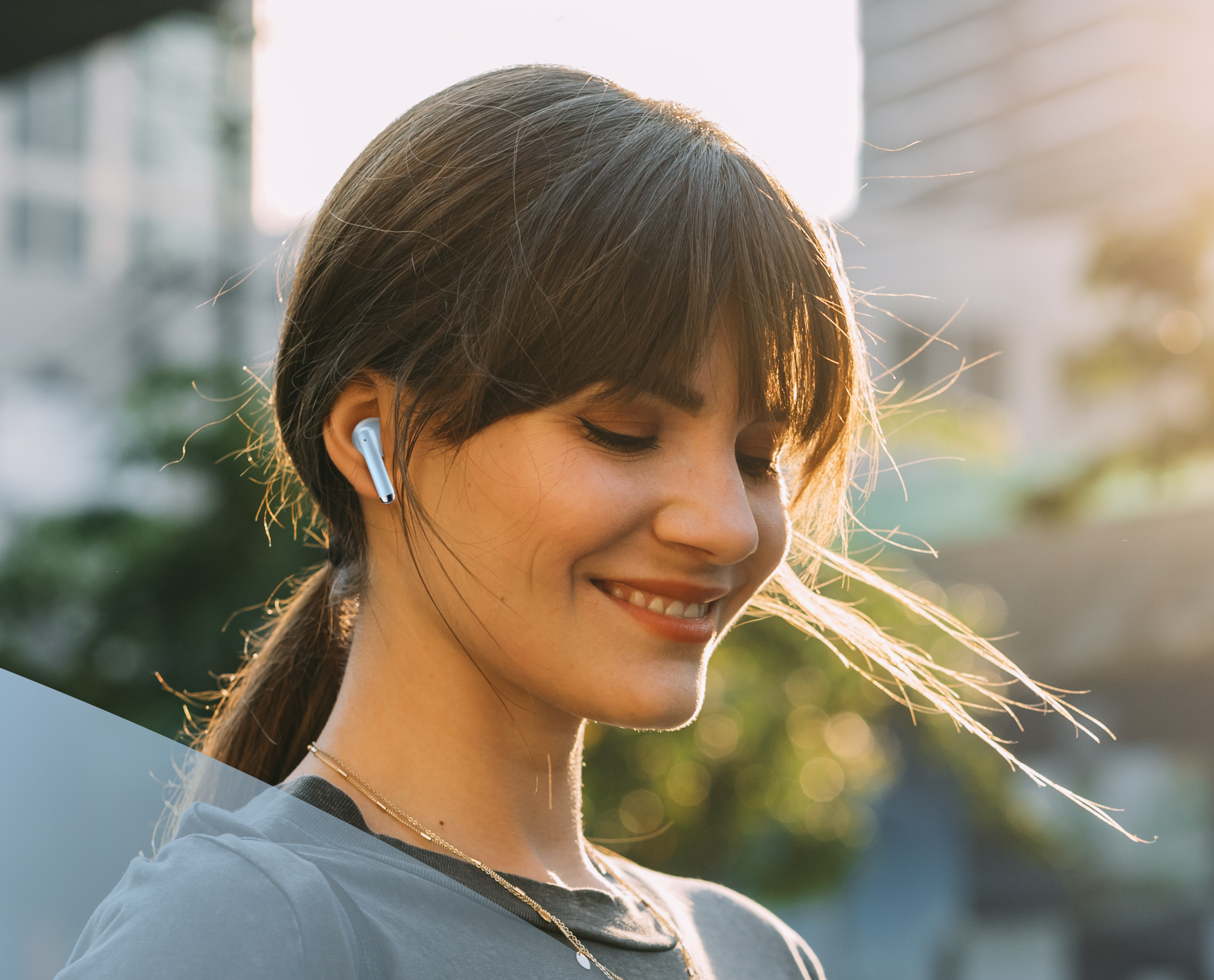 Uśmiechnięta kobieta w jasnoniebieskich słuchawkach. Jest na zewnątrz w słońcu. Włosy w kucyku.