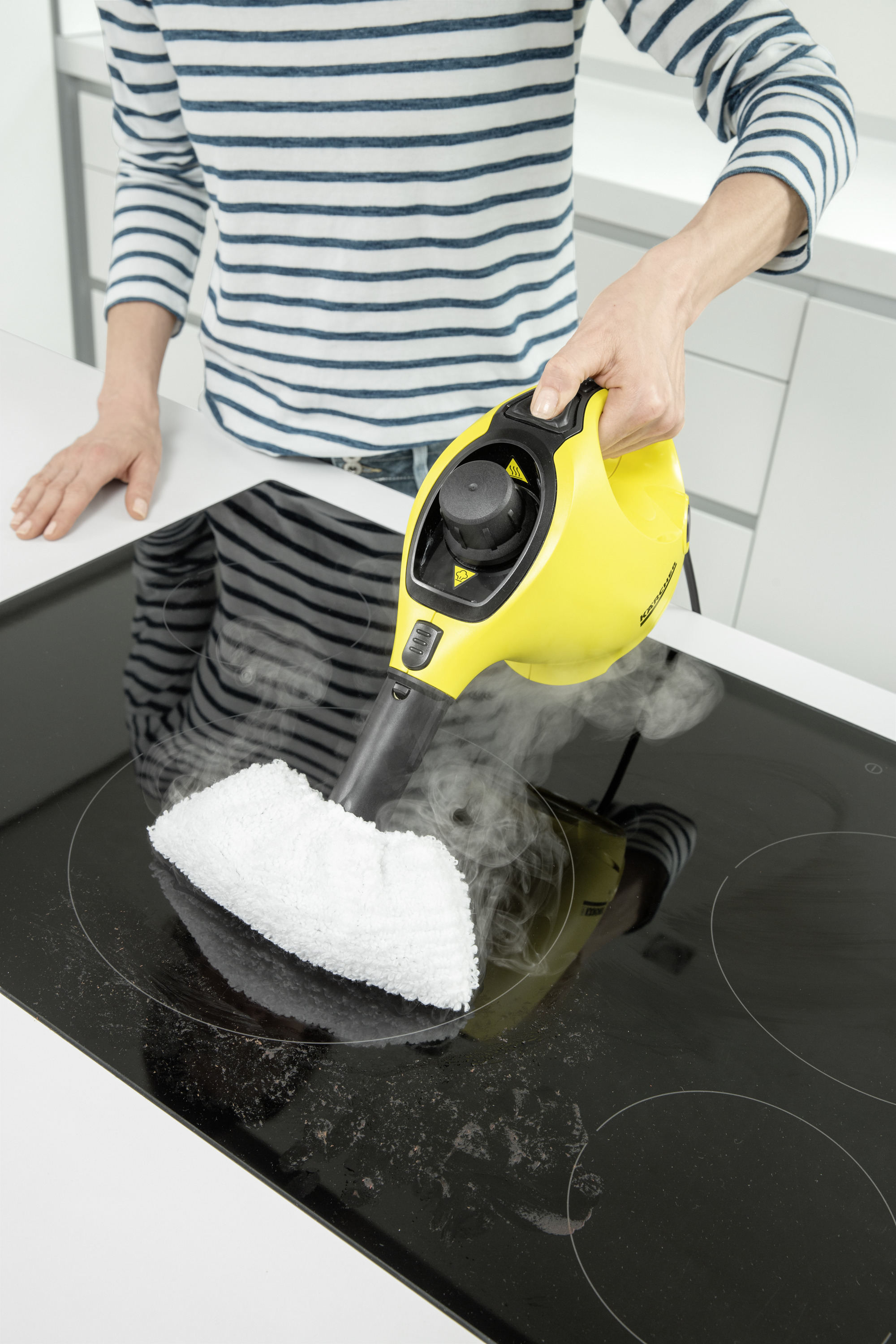 Una persona pulisce un piano cottura in vetro nero con un pulitore a vapore giallo, con vapore che sale.