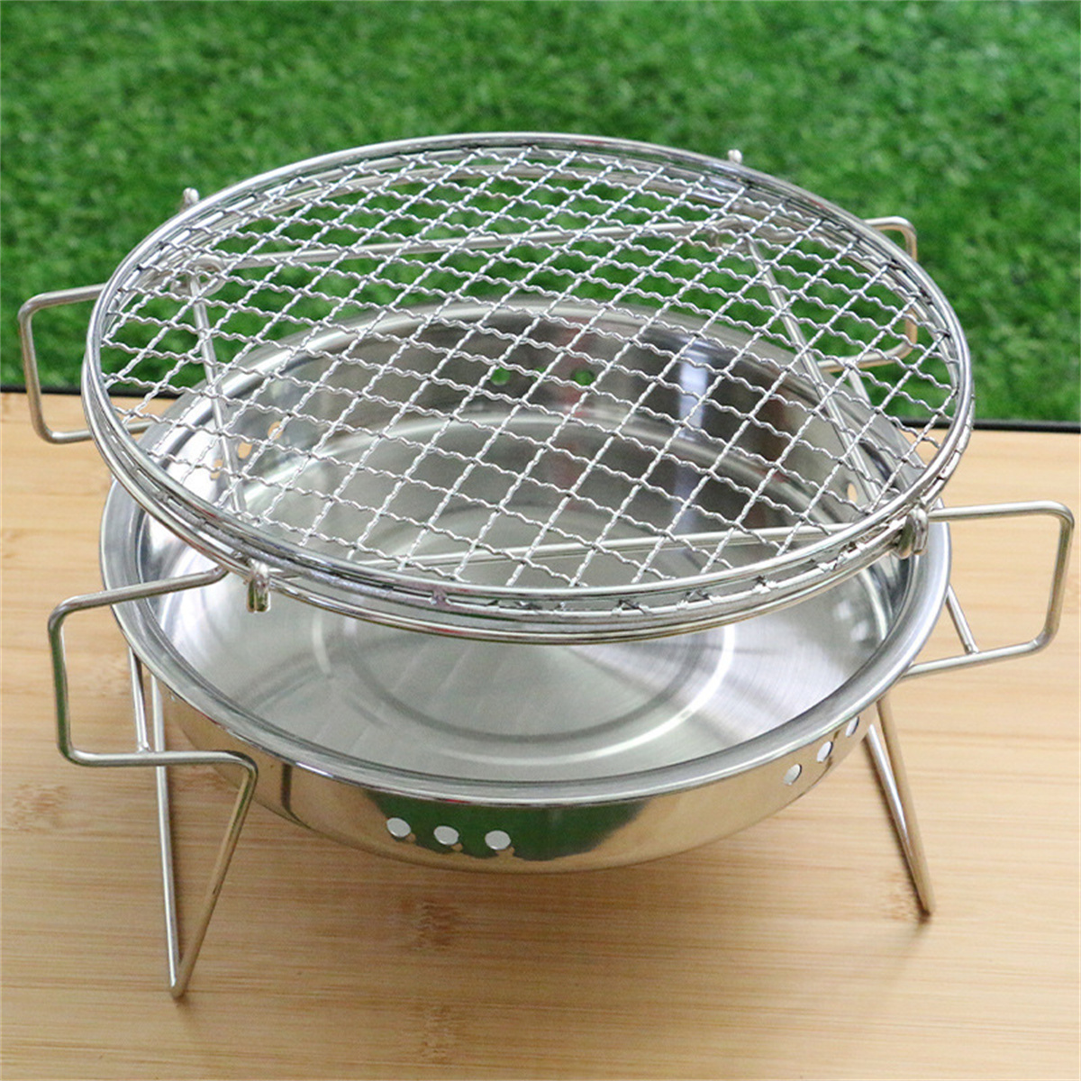 Ein Edelstahlgrill mit kreisförmigem Design. Auf Holzoberfläche platziert. Grünes Gras im Hintergrund.