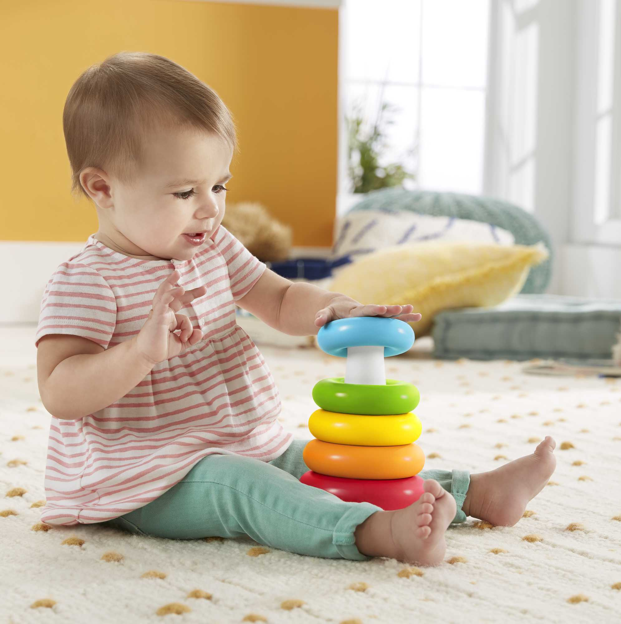 Baby sitzt auf einem gemusterten Teppich, spielt mit bunten Stapelringen, lächelt.
