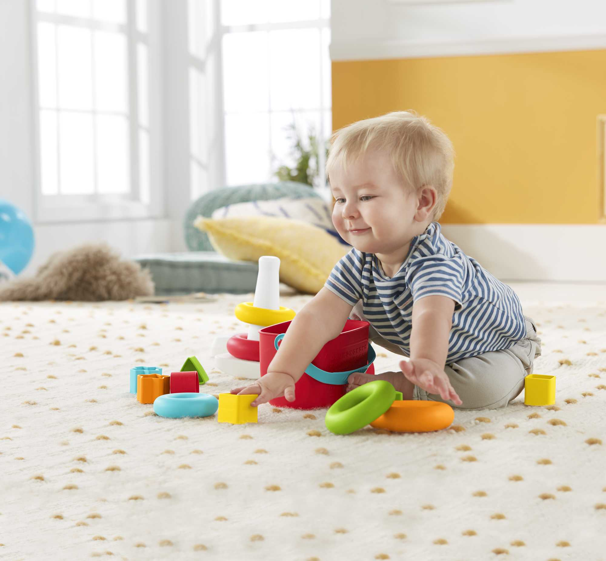 Baby spielt mit bunten Stapelringen und Blöcken auf einem gemusterten Teppich, lächelt glücklich.