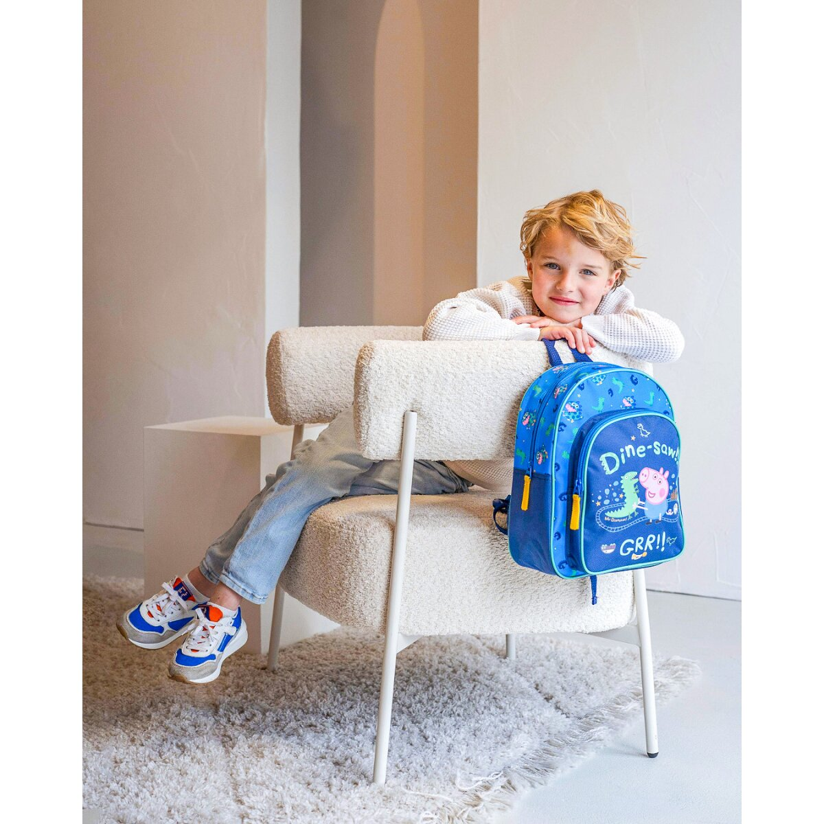 Junge mit Rucksack sitzt auf einem Stuhl. Weißes Zimmer mit Teppich und Stuhl.