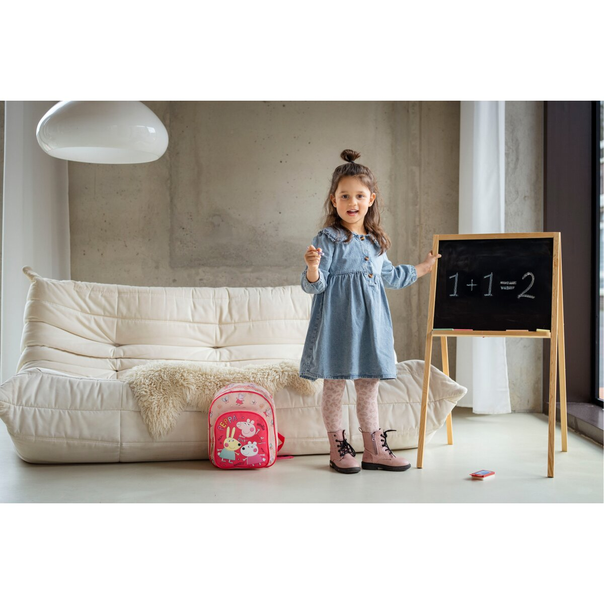 Mädchen steht an einer Tafel mit 1+1=2. Rucksack und Couch sind in der Nähe.