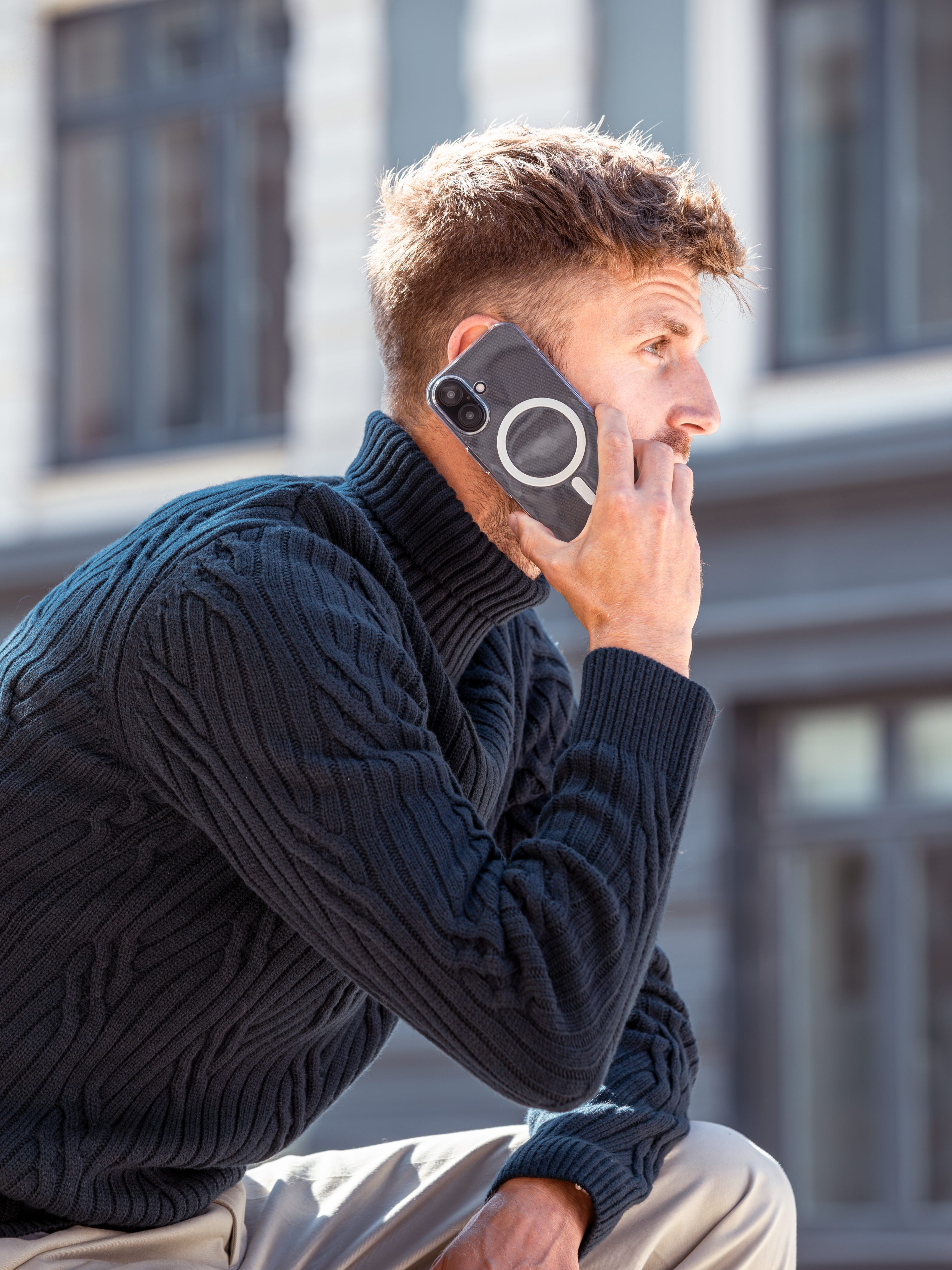 Un uomo parla al telefono con una custodia trasparente. Indossa un maglione blu scuro, con uno sfondo all'aperto.