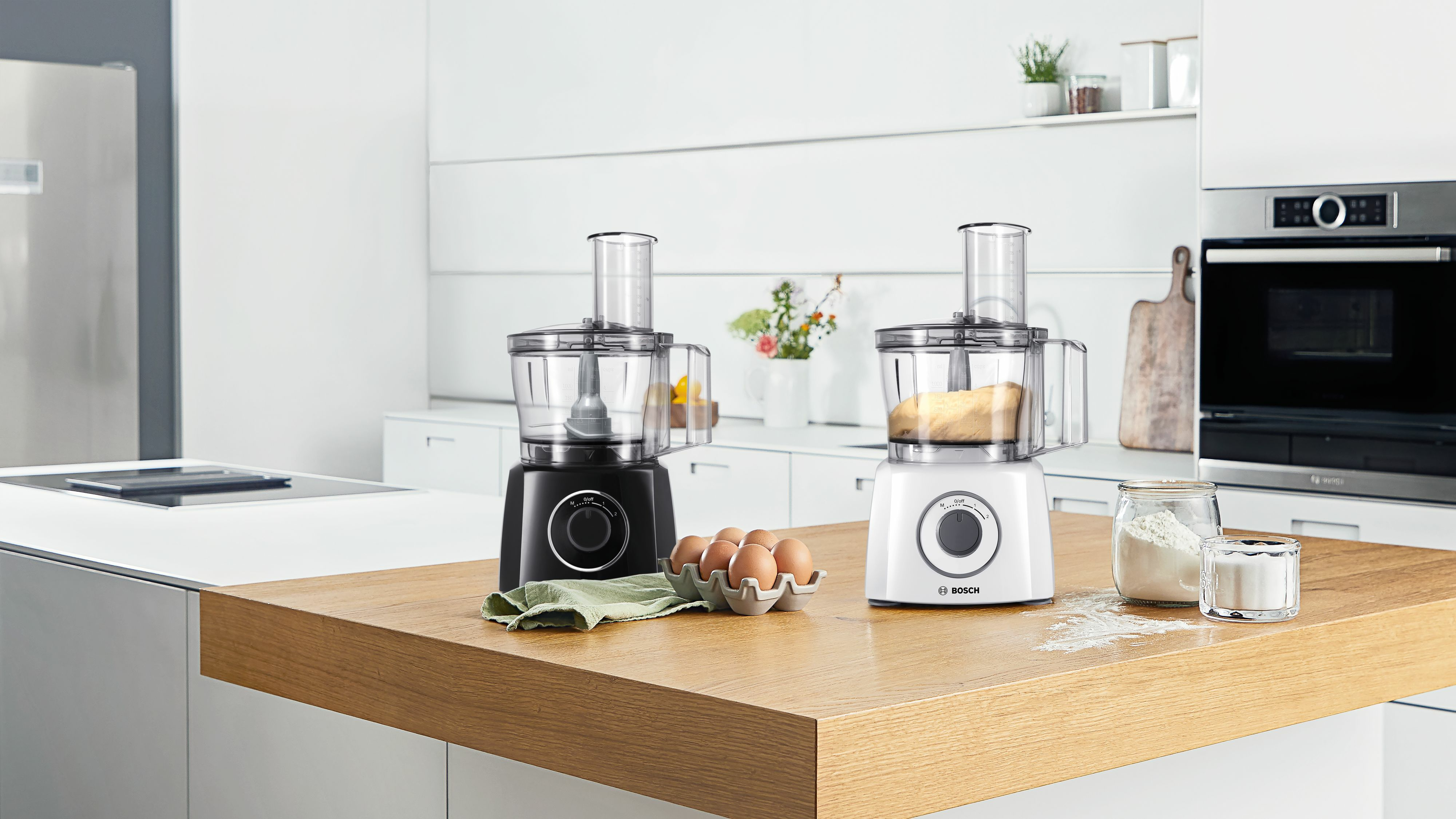 Dwa roboty kuchenne, czarny i biały, ze składnikami na drewnianej powierzchni w nowoczesnej kuchni.