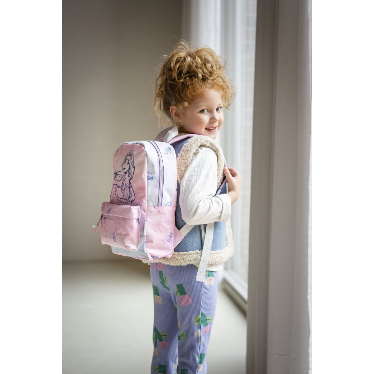 Ein junges Mädchen mit einem Rucksack mit Elsa-Grafik, steht in der Nähe eines Fensters und lächelt.