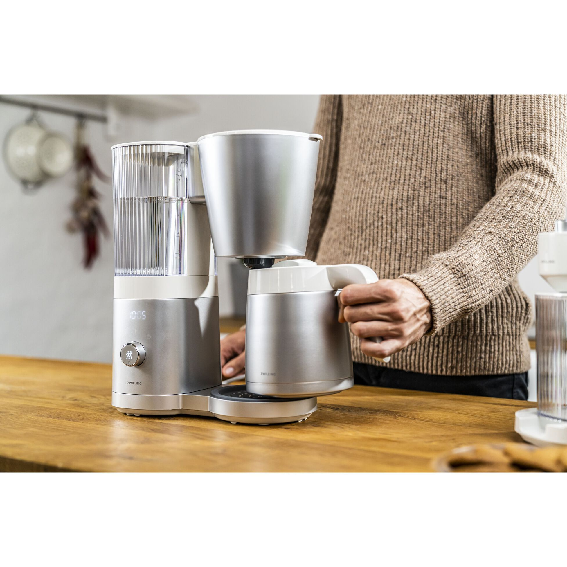 Persona sosteniendo una cafetera, cafetera sobre mesa de madera, cocina al fondo.
