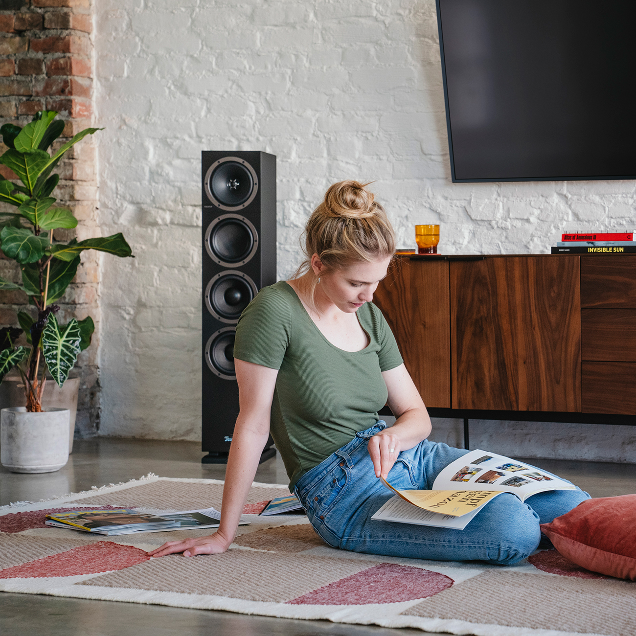 Frau liest Buch, sitzt auf Boden. Lautsprecher und TV im Hintergrund.