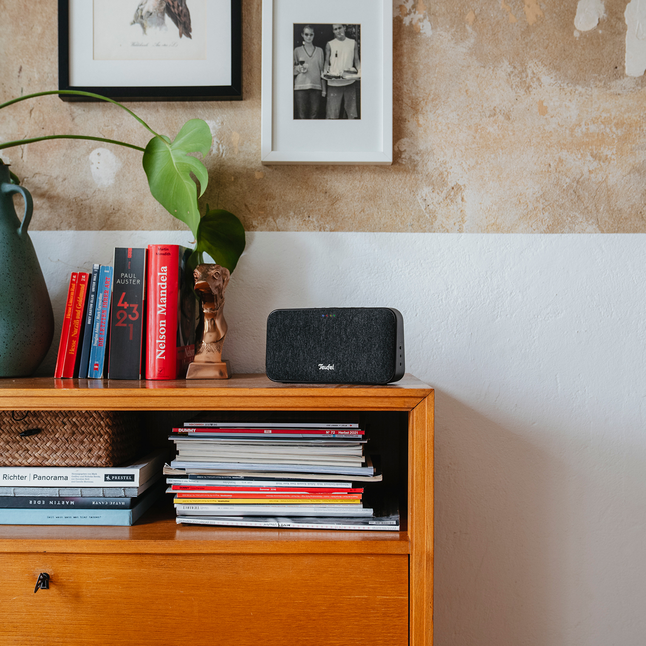 Luidspreker op een houten dressoir met boeken, een vaas en ingelijste foto's. De muur is geverfd.